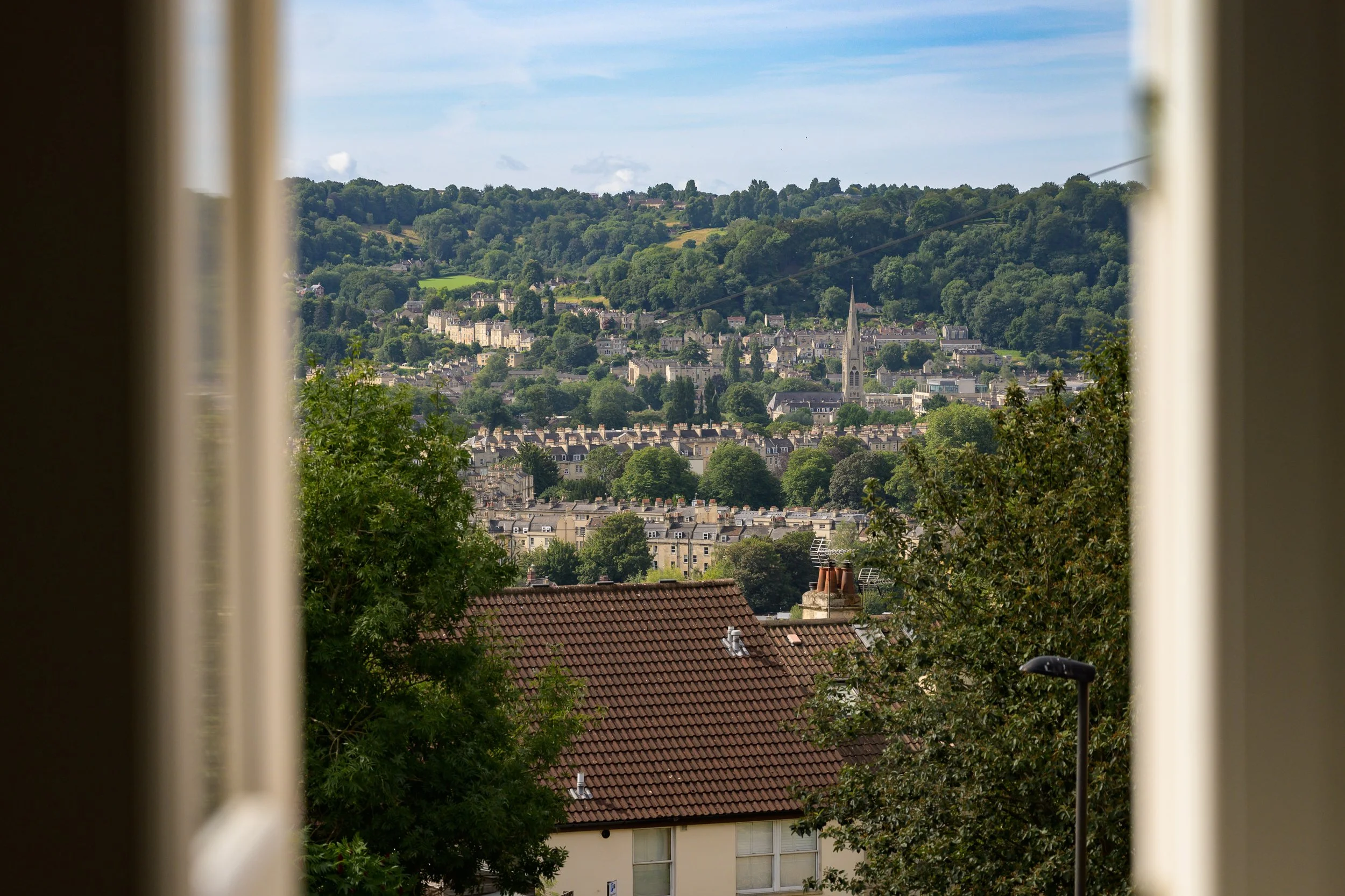 View of a hillside of city of Bath with buildings, trees, a church steeple, and green landscape, seen through a window frame.
