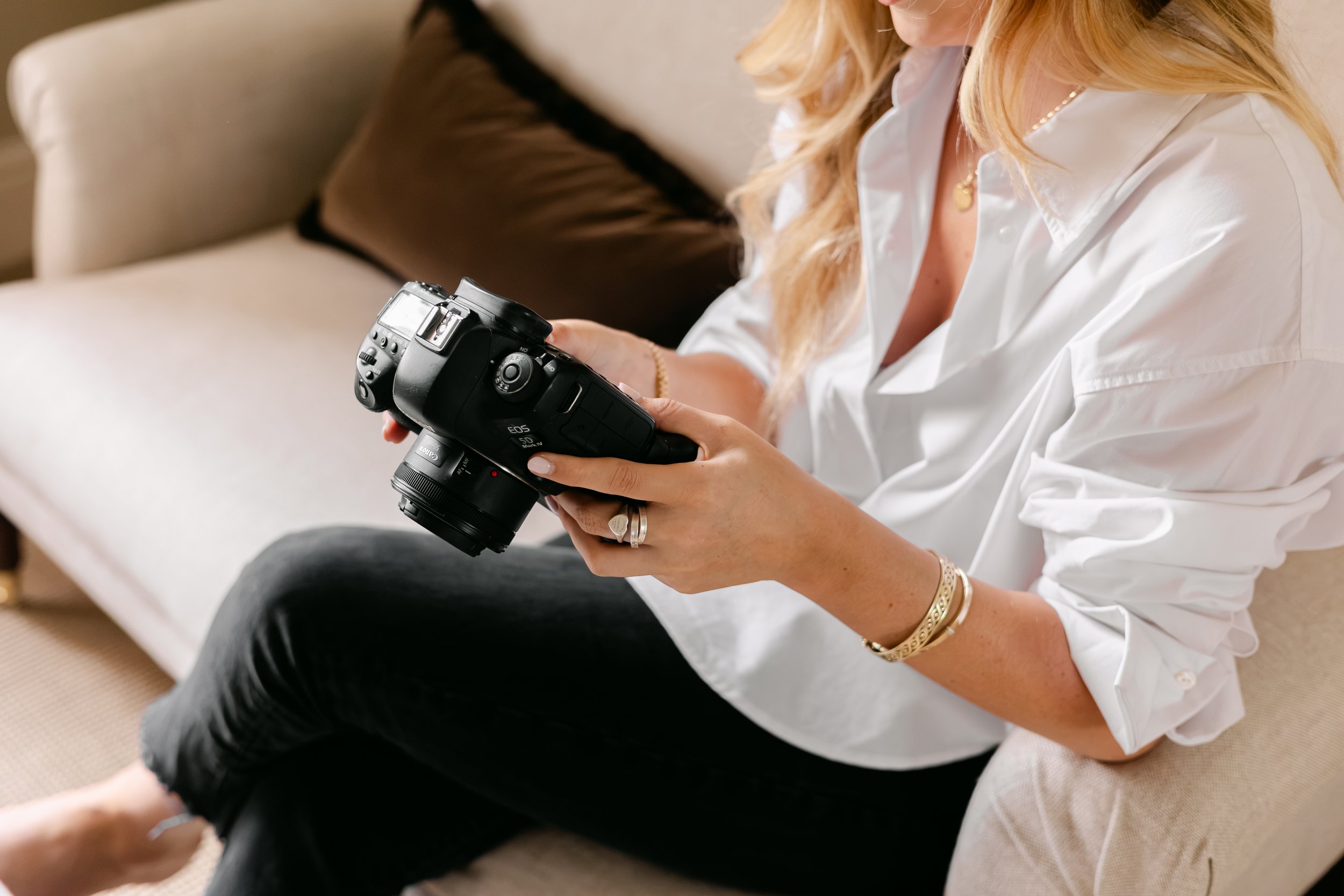 A woman with blonde hair sitting on a beige couch, holding a professional camera in her hands, wearing a white shirt and black pants, with jewelry including rings, bracelets, and a necklace.