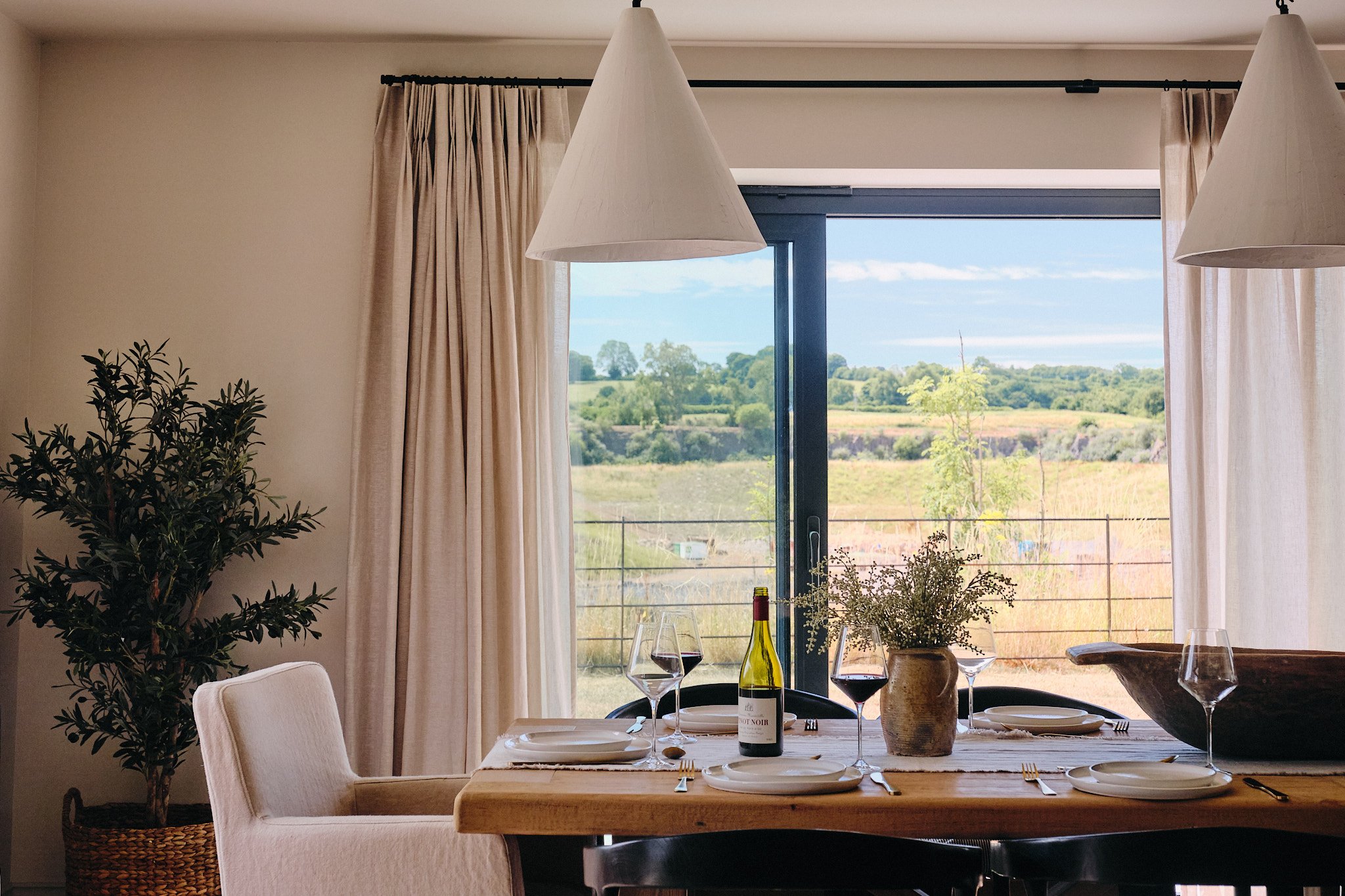 A dining table set with white plates, wine glasses, and a bottle of red wine in a room with large windows and cream curtains, overlooking a green outdoor landscape with trees and a cloudy sky.