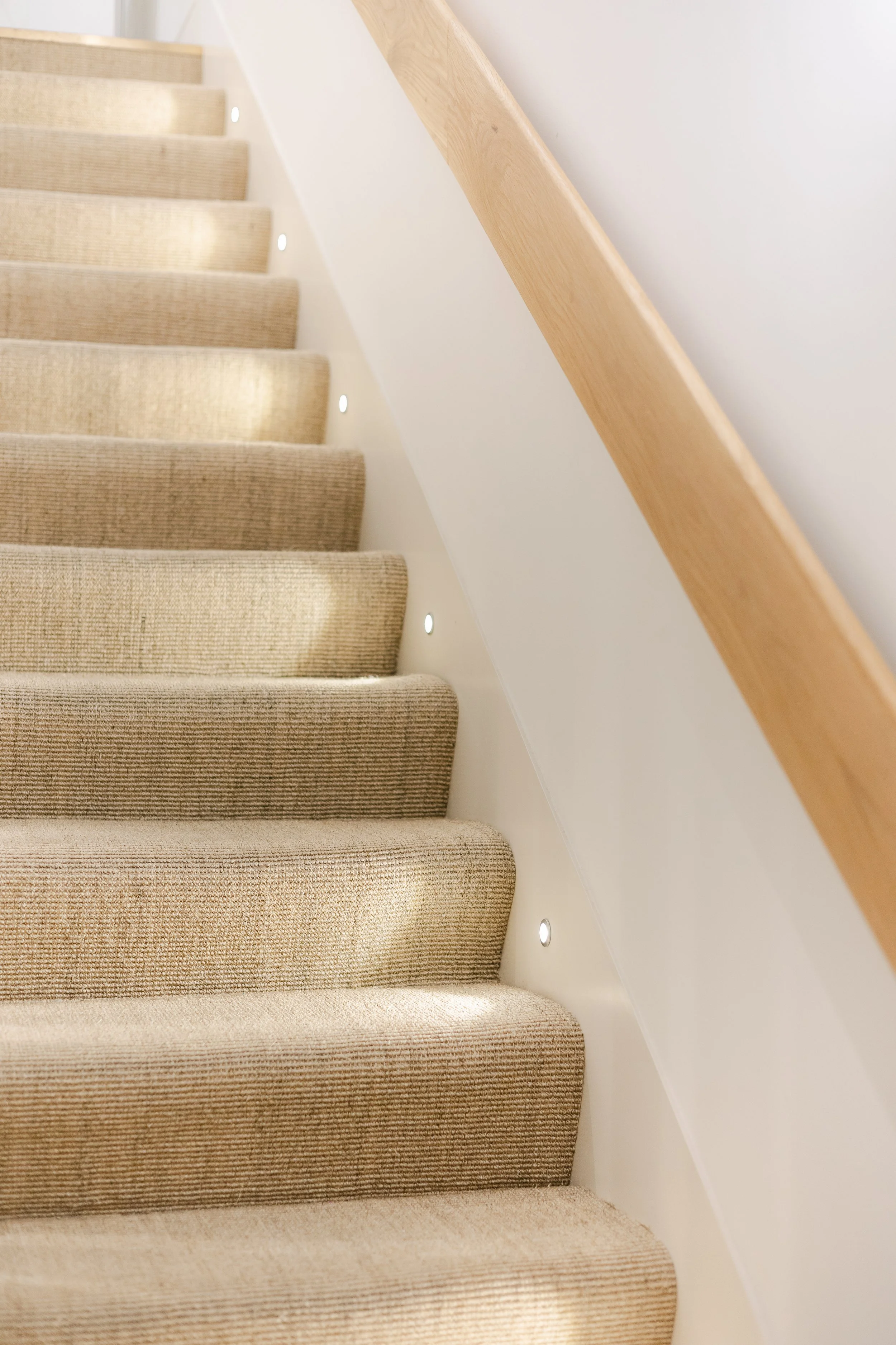 Indoor staircase with beige carpeted steps, wooden handrail, and small built-in lights along the side.