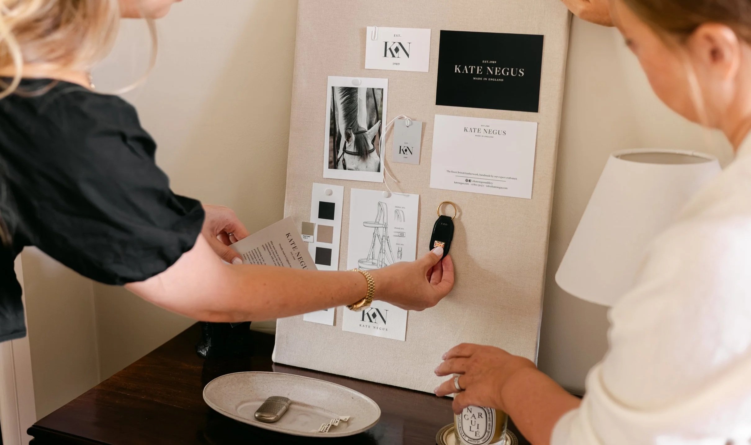 Two women are discussing branding materials for Kate Negus, a British leatherwork company. They are examining a display board with branding images, a photo of a horse, and design sketches, while one woman holds a key fob and a sheet of paper.