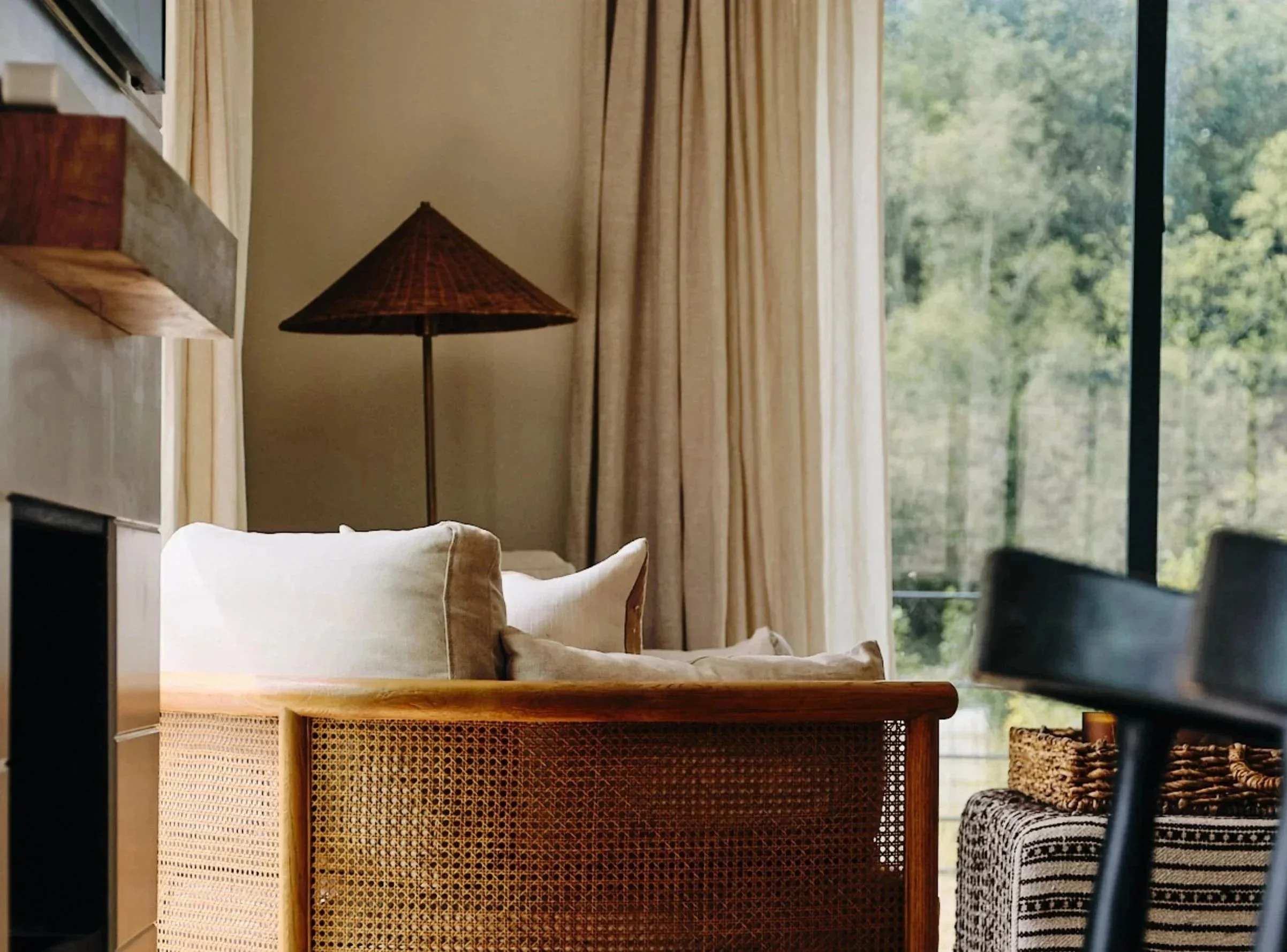 A cozy living room with a wicker sofa, beige cushions, a wooden side table, a tall floor lamp with a conical shade, and large windows with creamy curtains revealing greenery outside.