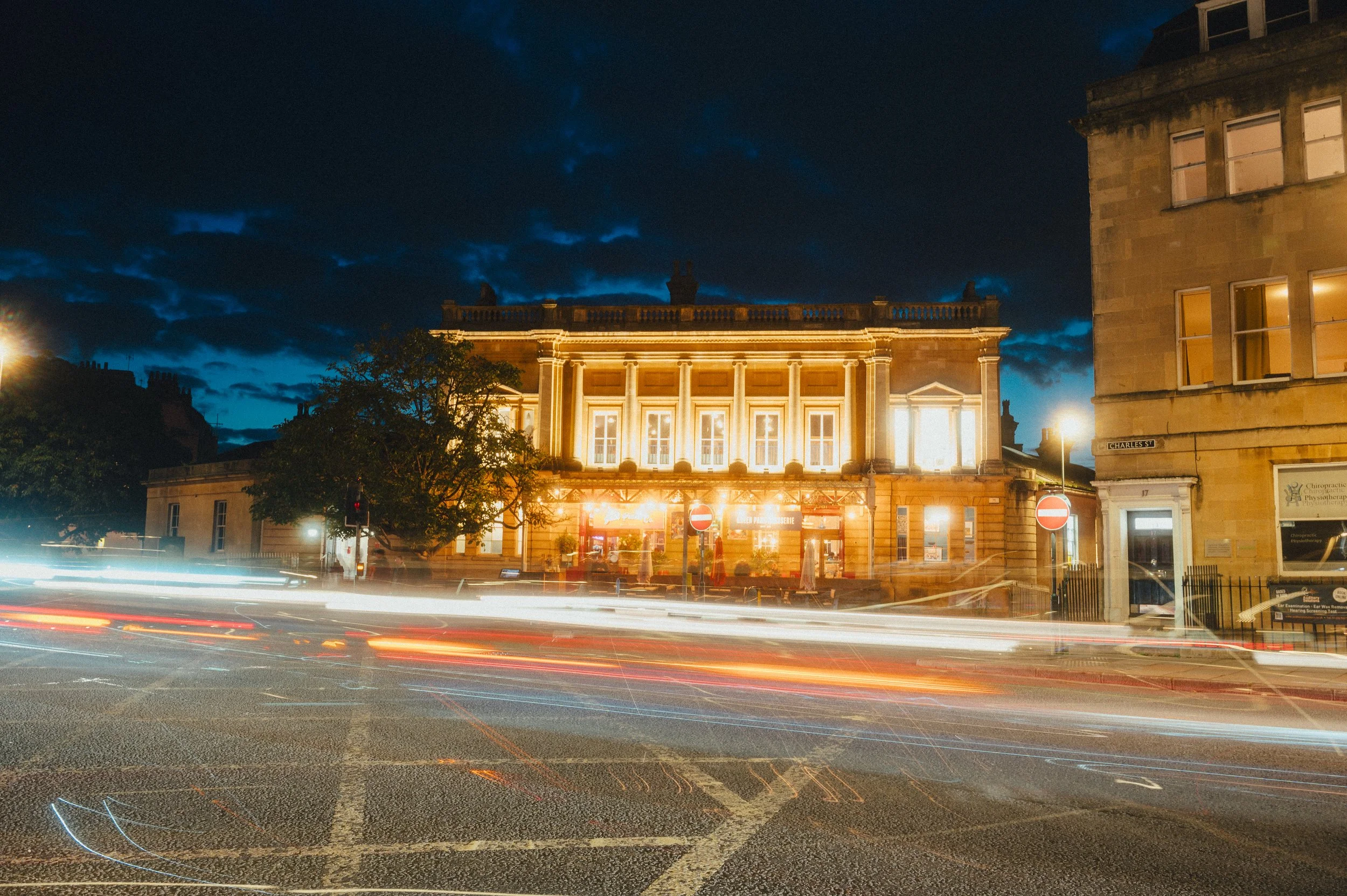 Nighttime city street with a historic building illuminated with yellow lights, light trails from moving cars, dark clouds in the sky, and street signs, including no entry signs.