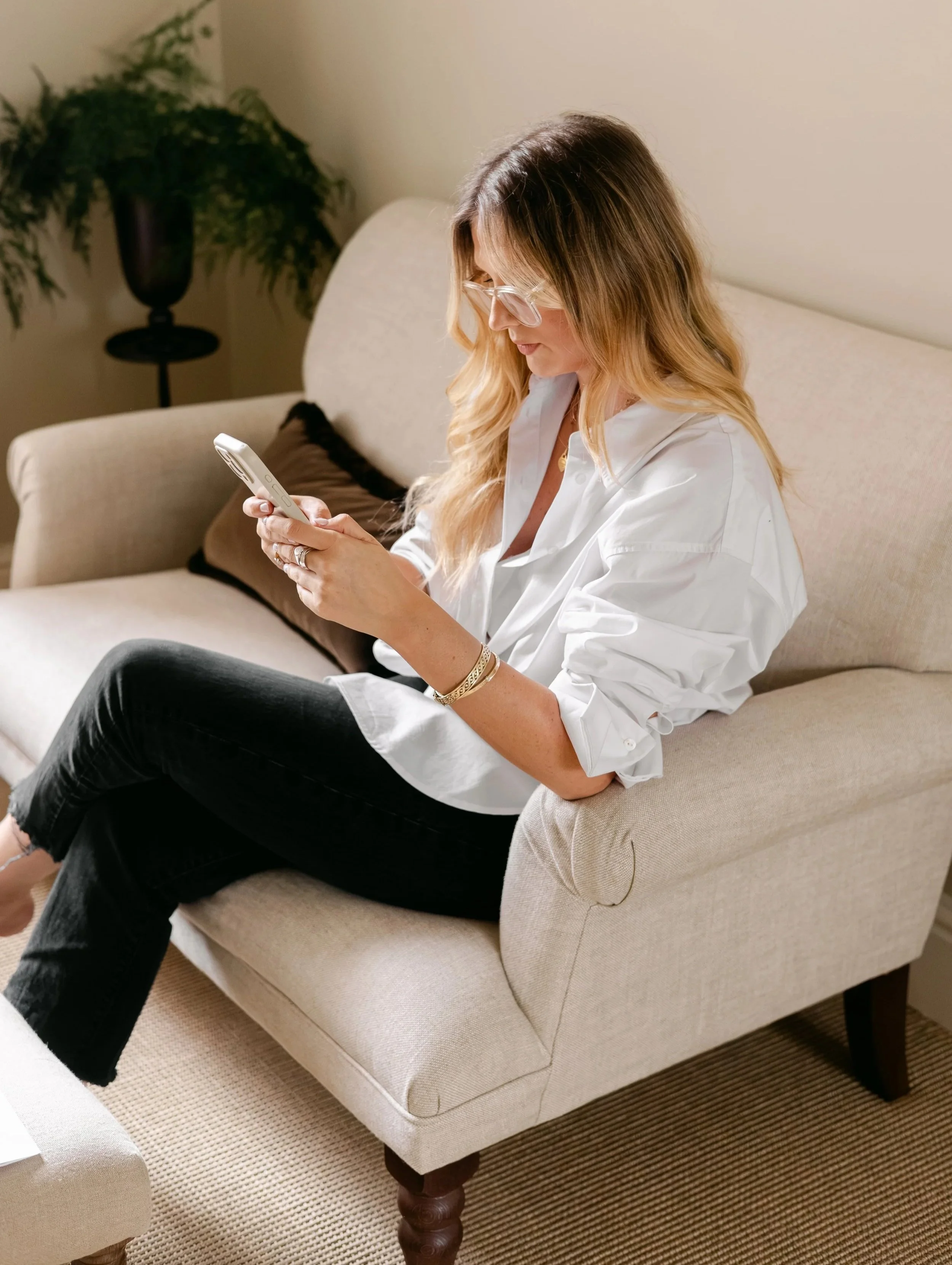 A woman with long, wavy hair wearing glasses and a white shirt, sitting on a beige sofa and looking at her phone, working on social media.