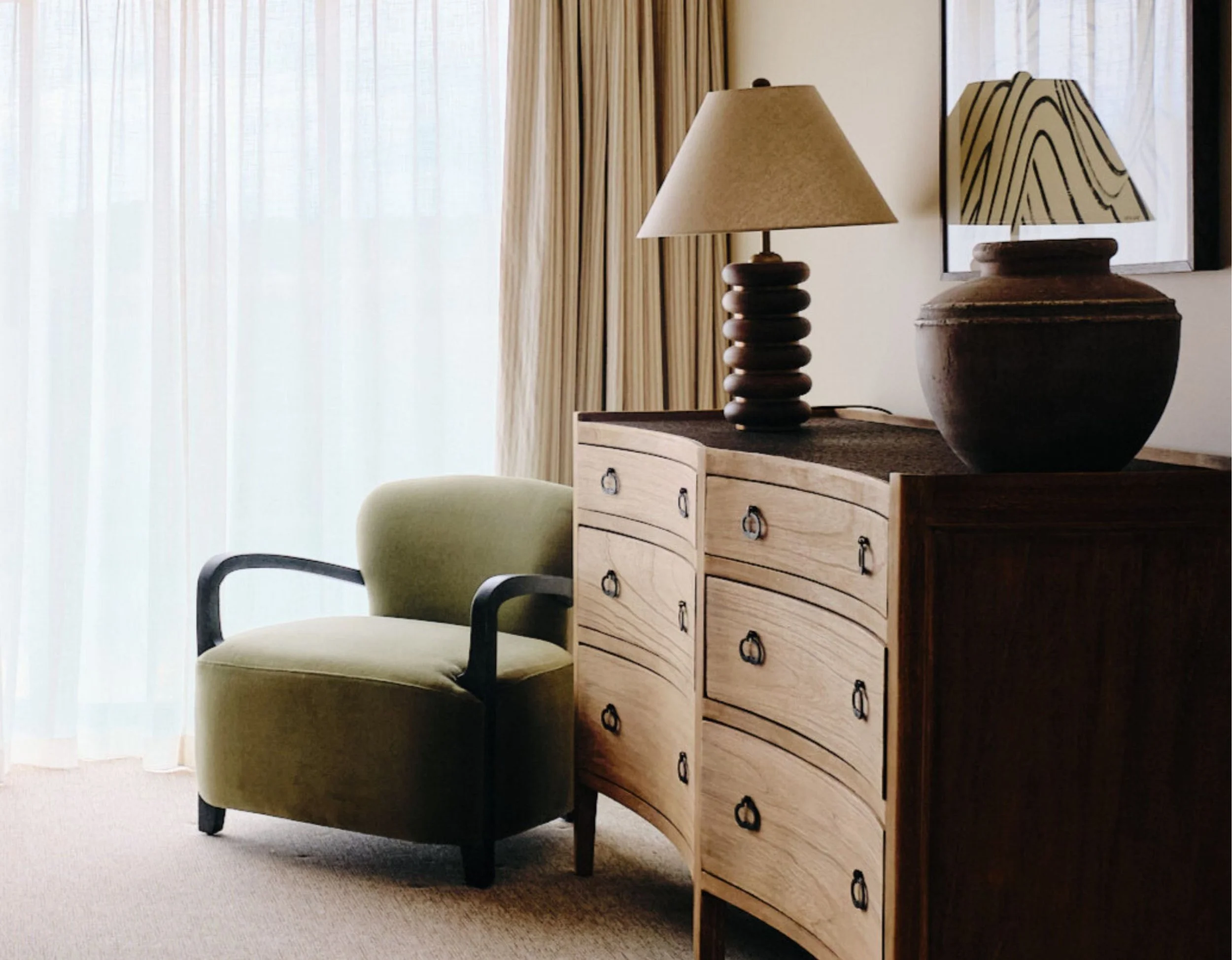 Luxurious hotel room with a pale green armchair, wooden dresser with black metal handles, and matching table lamps, near window with sheer curtains.