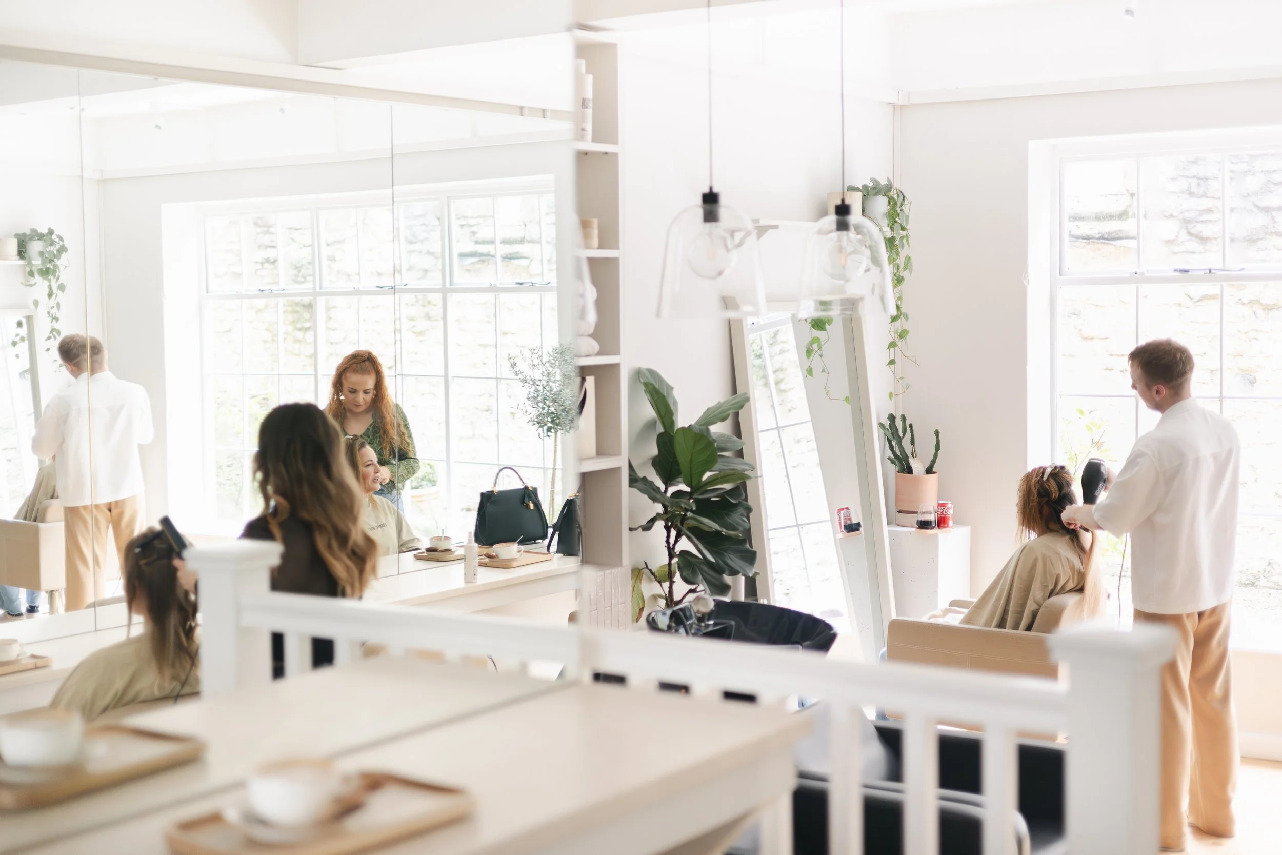 A hairstylist blow-drying a woman's hair in a bright, modern salon with large windows and minimalistic decor.