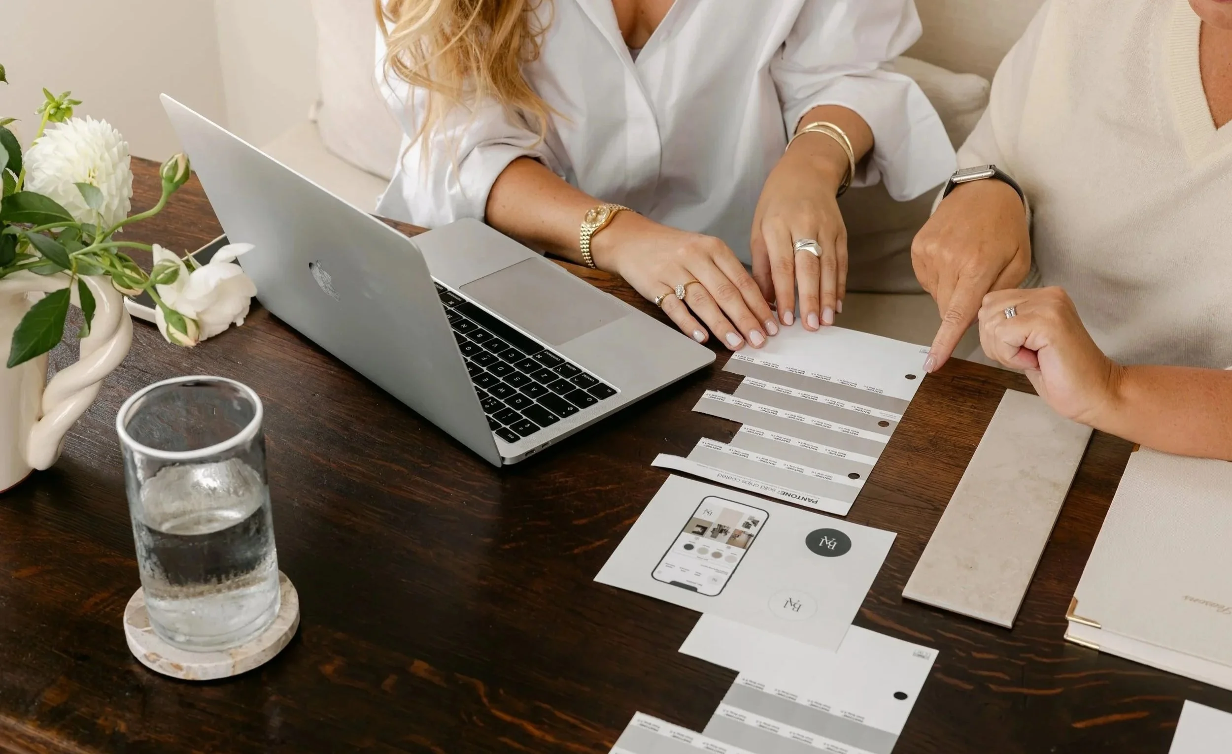 Two women are sitting at a wooden table looking at and discussing printed materials, with a silver laptop open in front of them. The table also has a clear glass of water, a floral arrangement in a white vase, and some promotional brochures or magazine pages. One woman is pointing at the printed materials. Both women are wearing jewelry, including rings, bracelets, and watches.