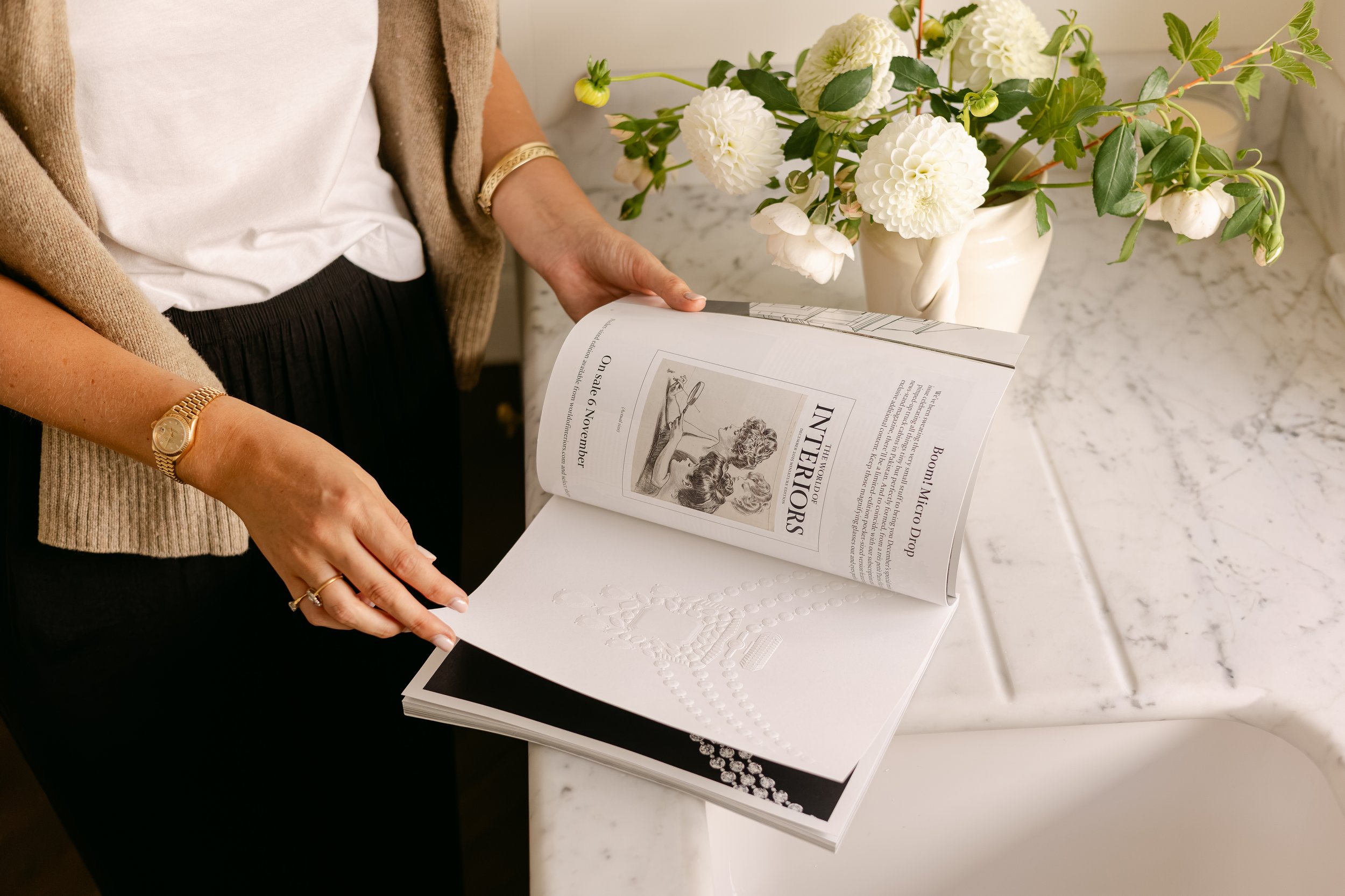 Person reading an Interior Design magazine with a garden scene on the cover, standing next to white flowers in a beige vase on a white marble countertop.