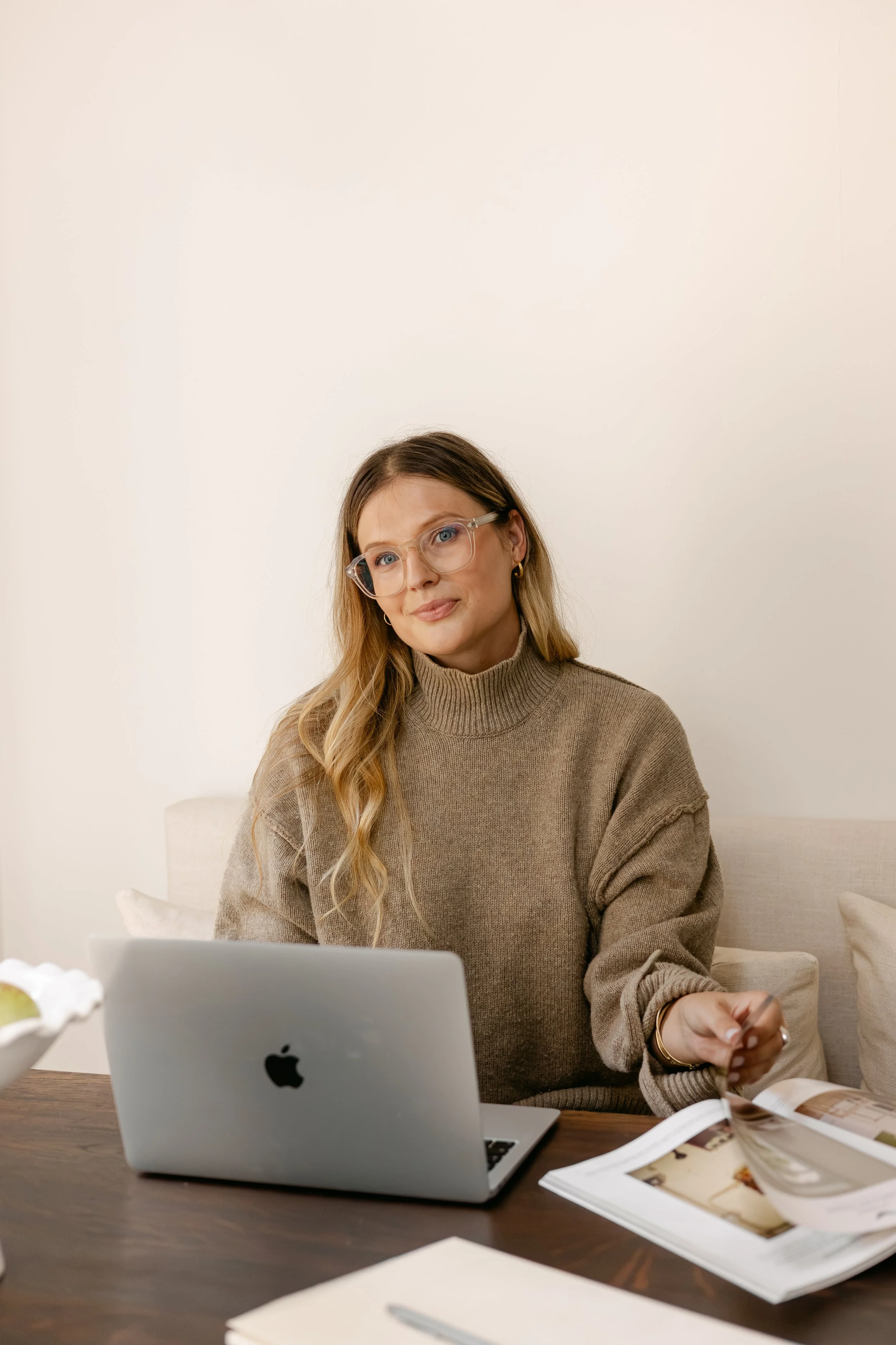 Founder of BA1 Studios behind a laptop with long blonde hair, wearing glasses and a brown sweater, surrounded by magazines and papers, in a interior designed room.