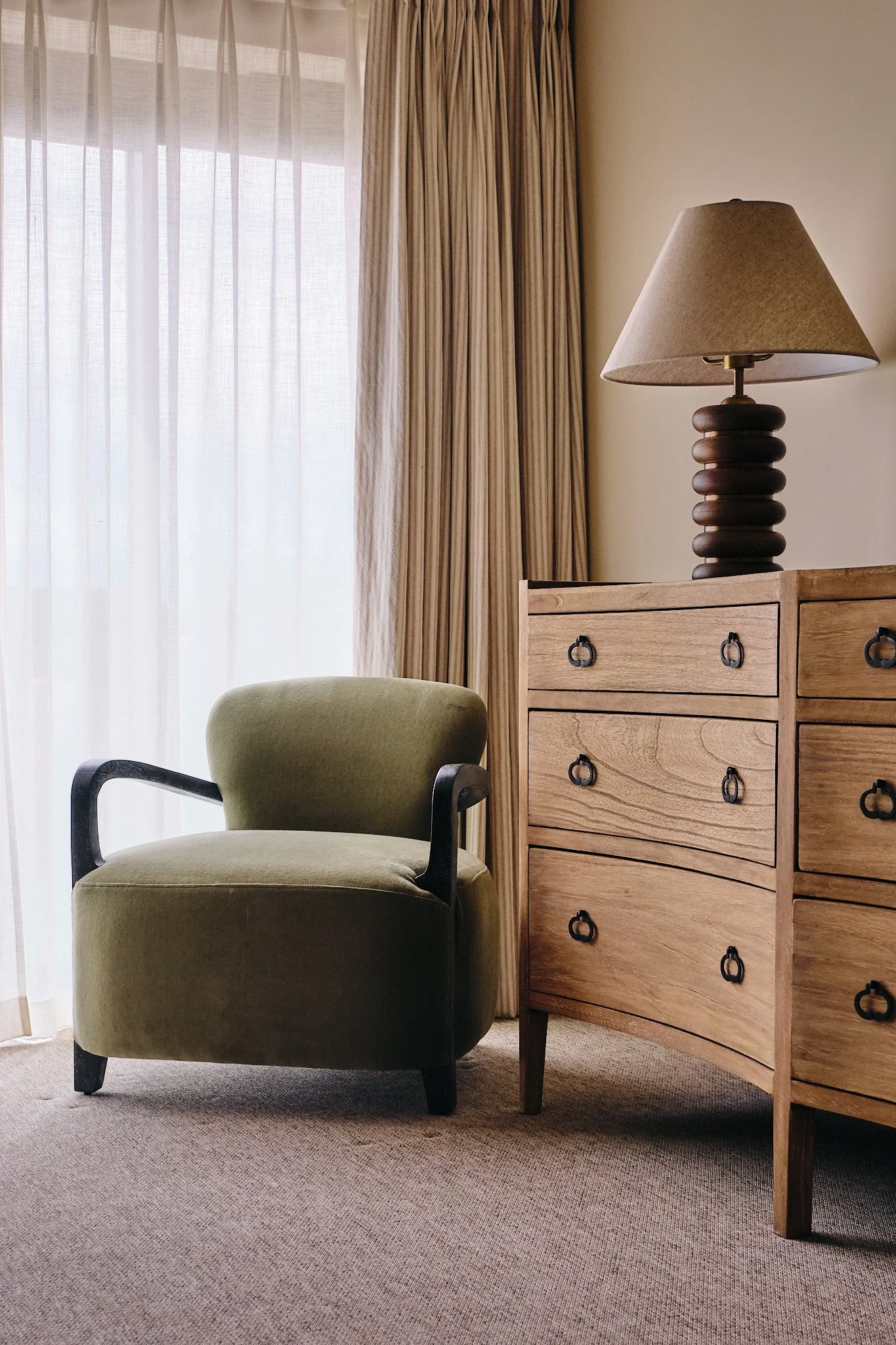 A living room corner with a green armchair, wooden dresser with drawers and a table lamp, beige curtains, and a sheer curtain covering a window.