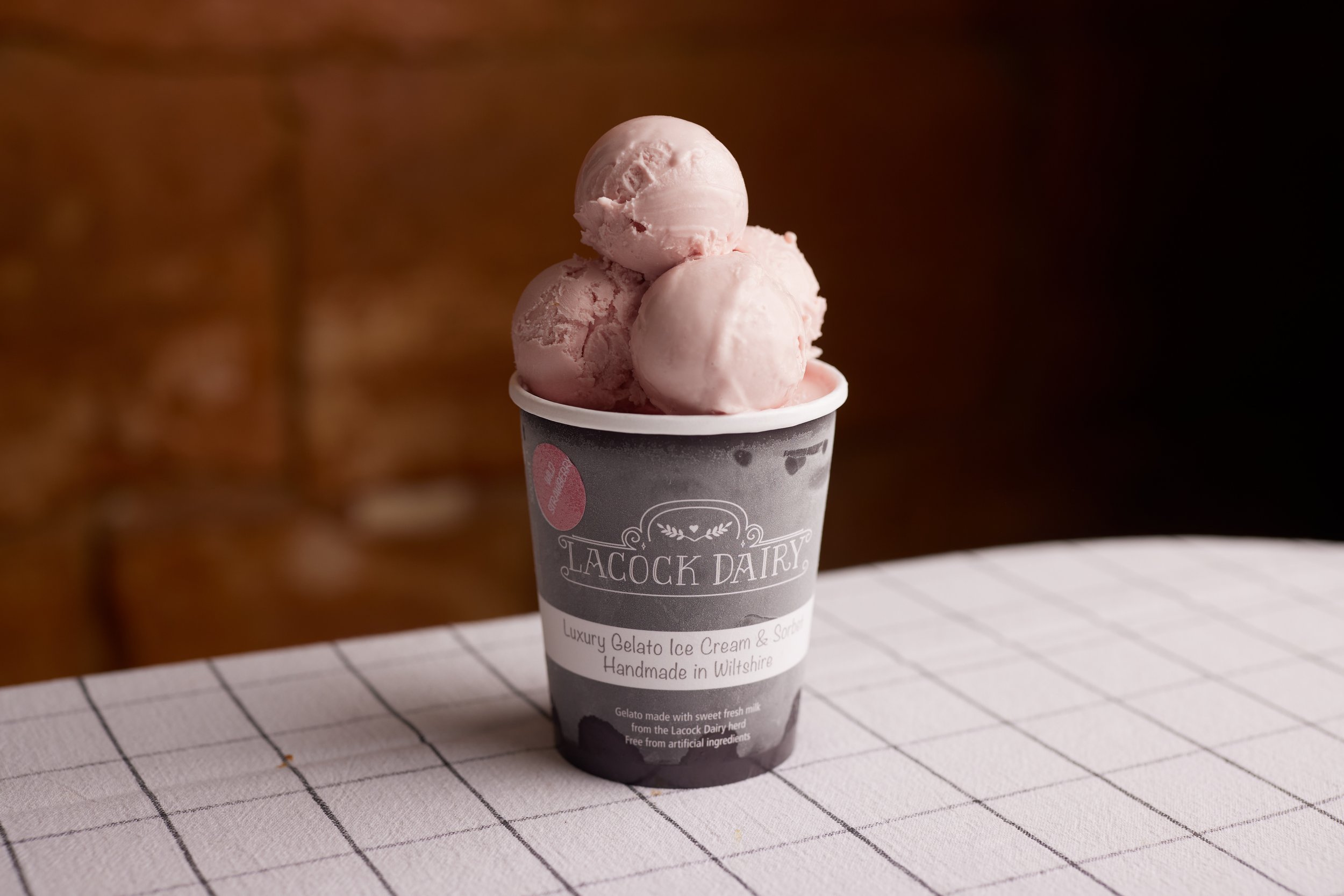 Three scoops of pink gelato in an ice cream cup on a checkered tablecloth.