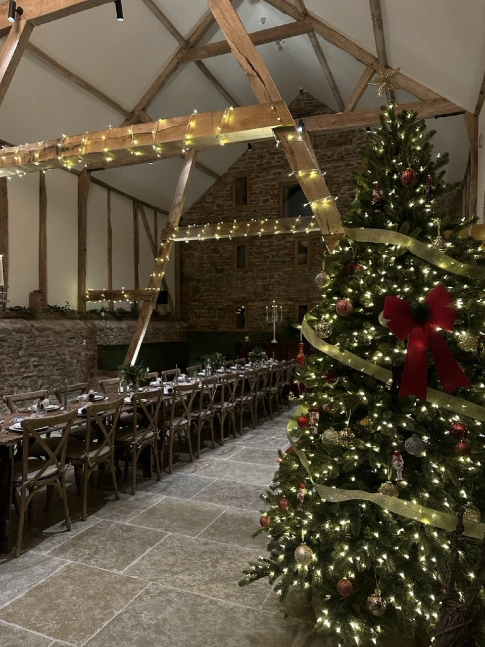 A decorated Christmas tree with lights, ornaments, a red bow, and gold ribbon in a rustic indoor space. A long dining table set for a meal is visible in the background, with chairs and place settings, under wooden beams with string lights.