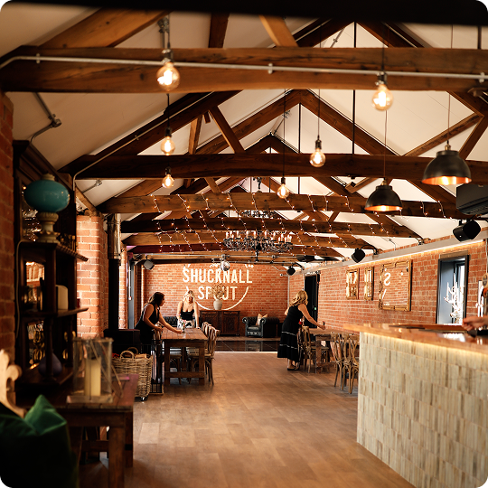 A wedding ceremony inside a rustic barn with exposed brick walls, wooden beams, and small windows, featuring a bride and groom exchanging vows in front of officiant, with guests seated on wooden chairs decorated with flowers.