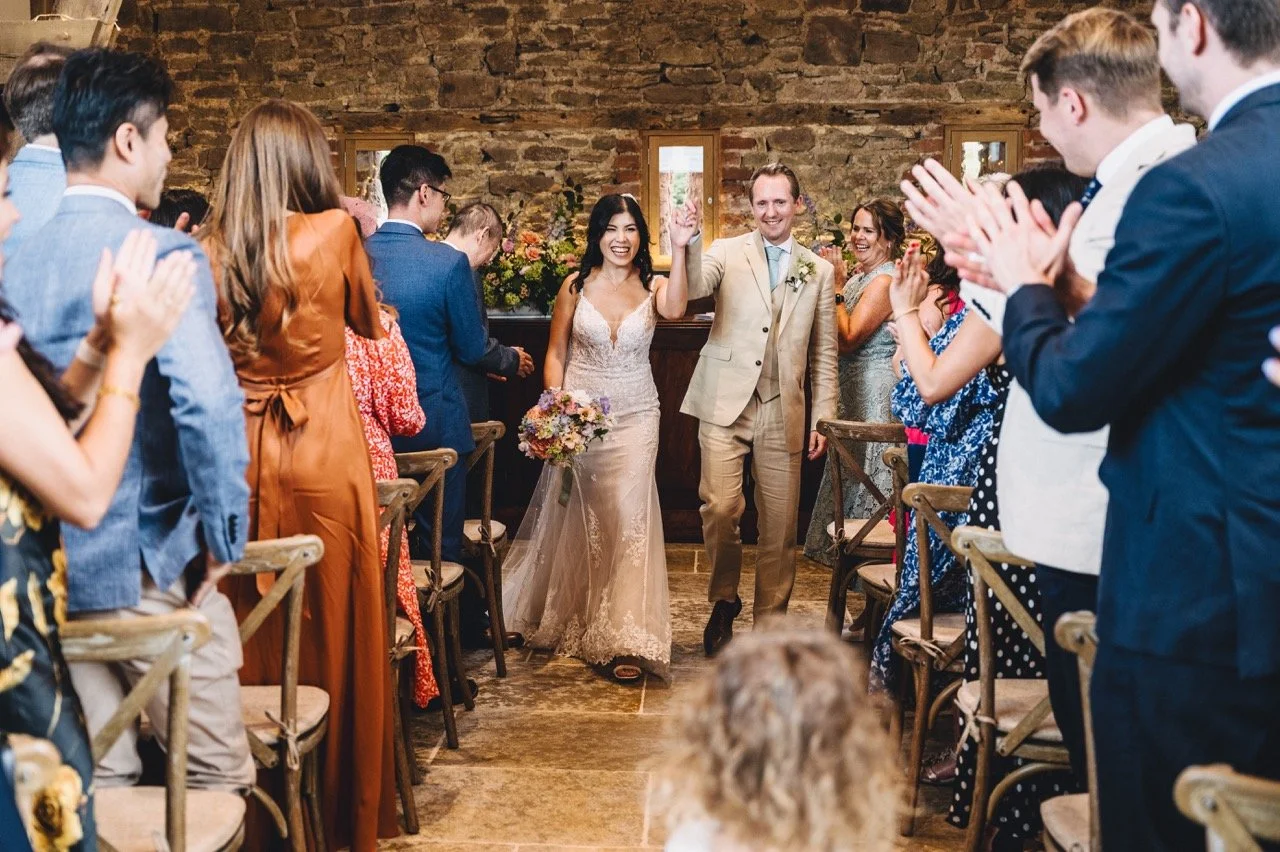 A wedding ceremony taking place in a rustic indoor setting with exposed brick walls and wooden beams, with guests seated on wooden chairs, and a bride and groom standing together exchanging vows.