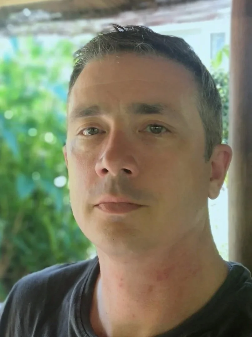 Close-up of a man with short dark hair, fair skin, and a slight smile, wearing a black shirt, with a blurred green outdoor background.