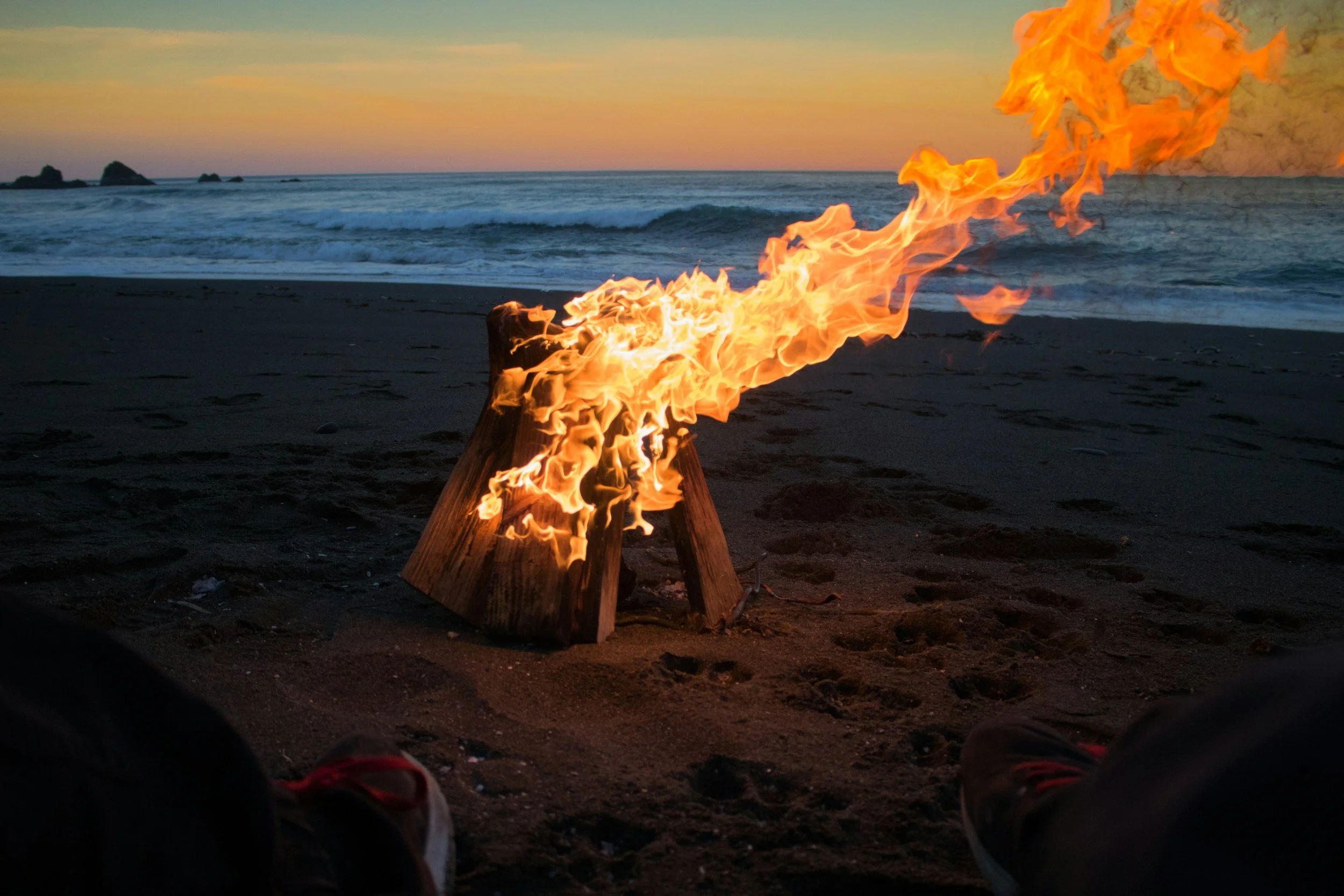 Campfire på stranden ved solnedgang med ocean i baggrunden og fødder i billedet