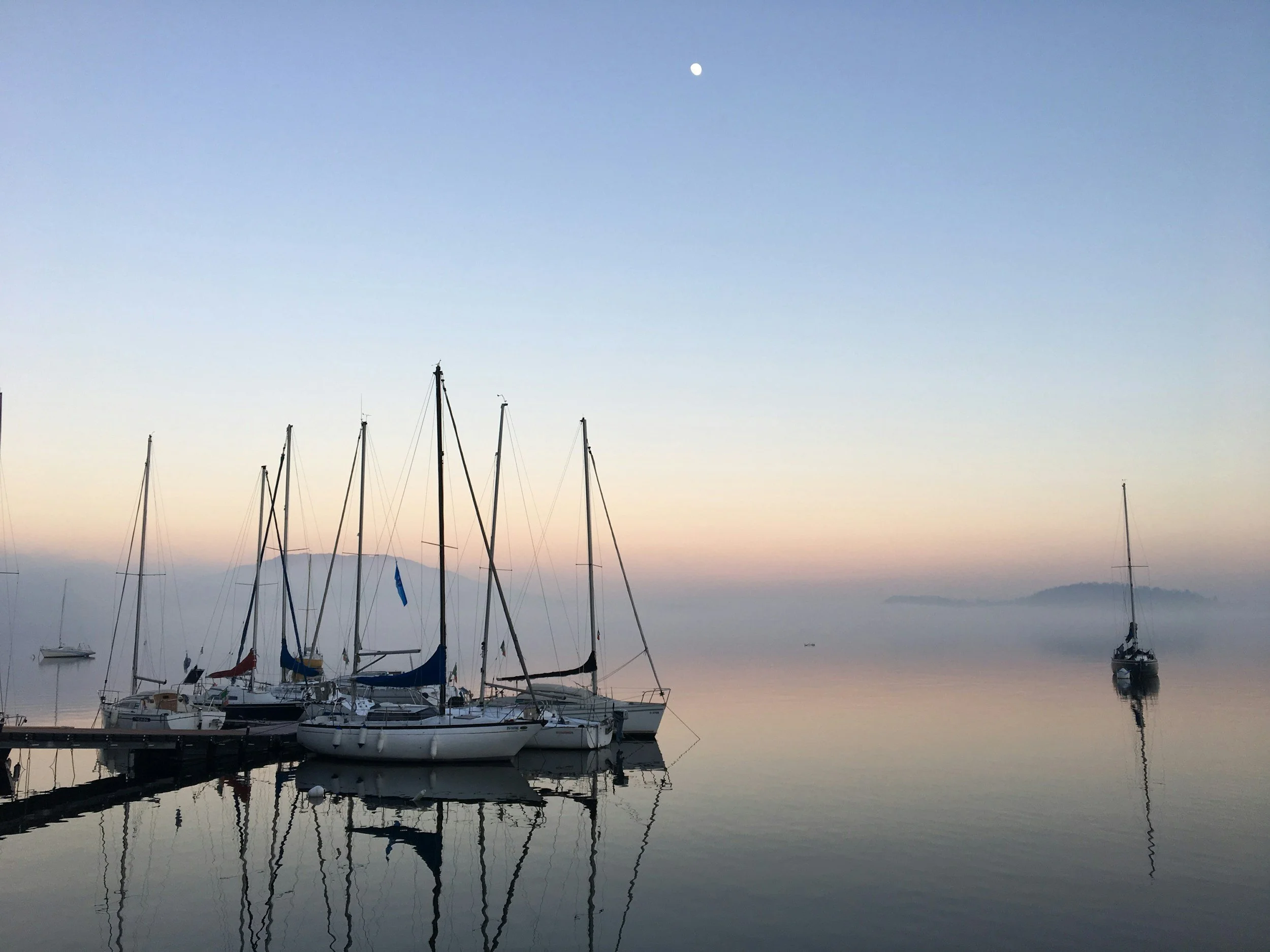 Både fortøjet i en rolig havn med spejlblankt vand, omkranset af let tåge og blidt pastelfarvet himmel ved solopgang eller solnedgang, med flere sejlbåde og en ø i baggrunden.