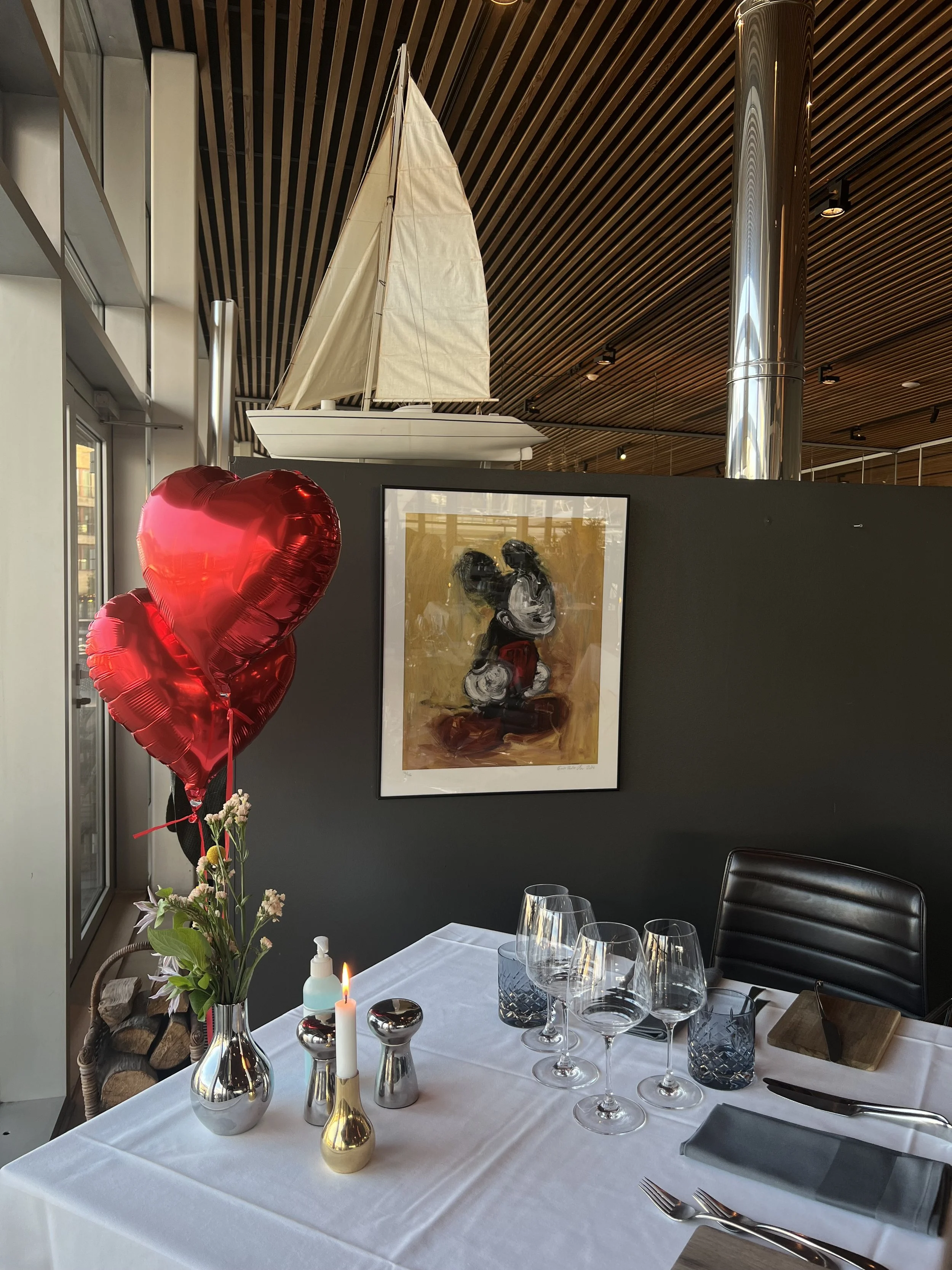 En borddækning med glasser, bestik, objekter, en lysestage, balloner og en vase med blomster, der er placeret ved vinduet i en restaurant med kunst på væggen og en modelsejlhel.