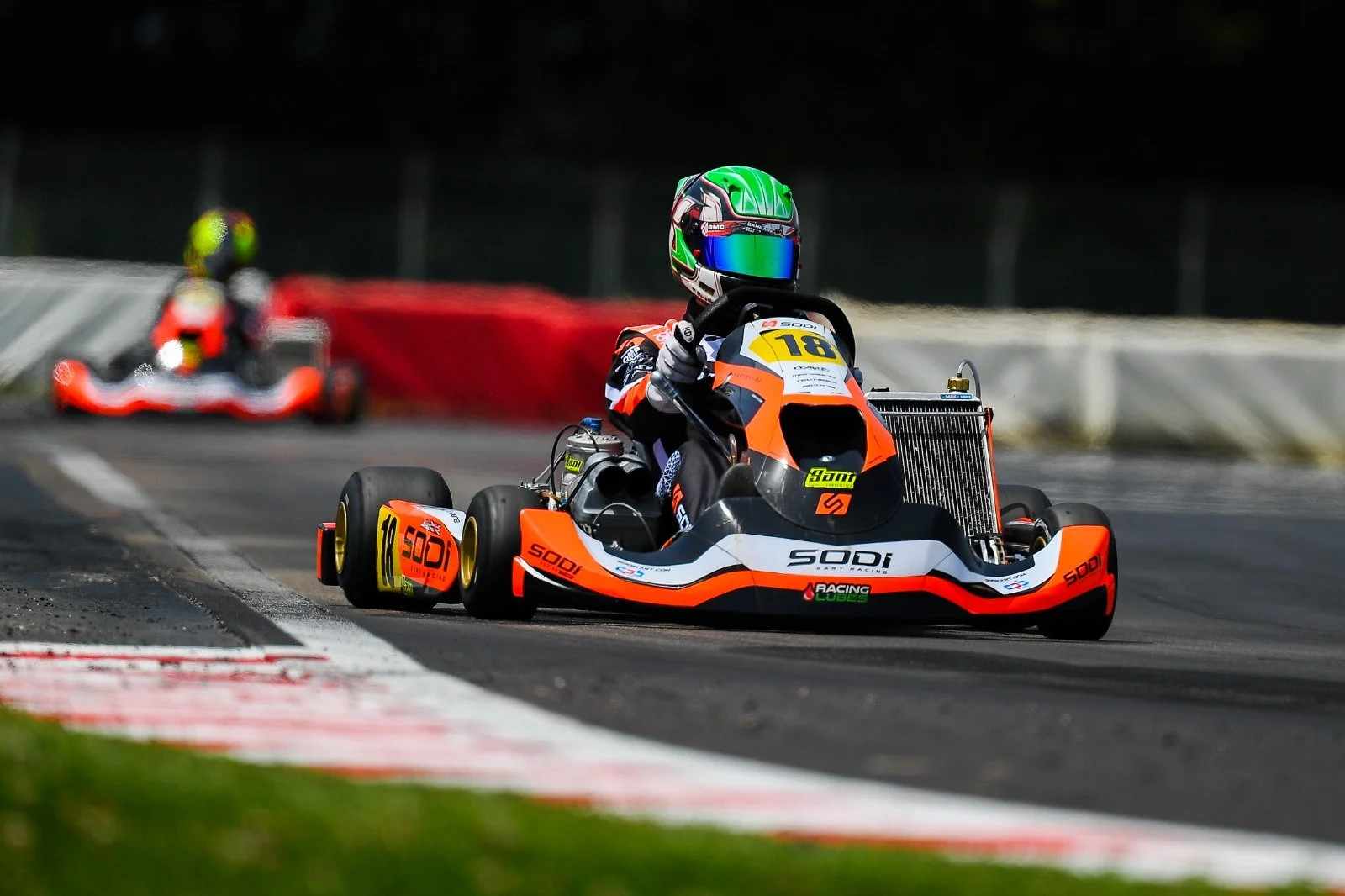 A go-kart driver wearing a green helmet, black racing suit, and gloves, leaning into a turn on a race track, with a blurred red and white barriers and another competitor in the background.