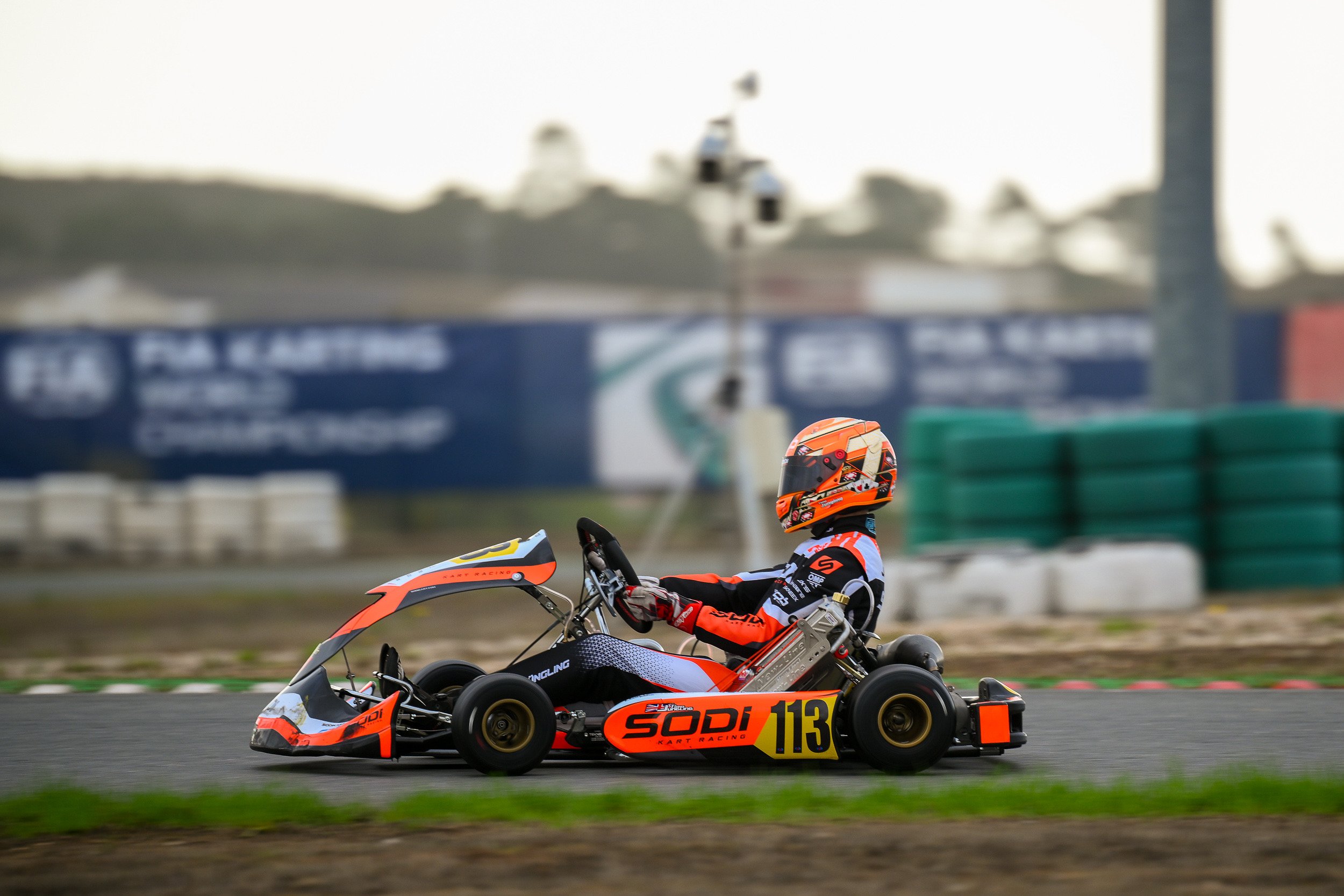 Go-kart racer wearing orange and black helmet and racing suit, driving a black and orange go-kart with the number 113 on a race track.