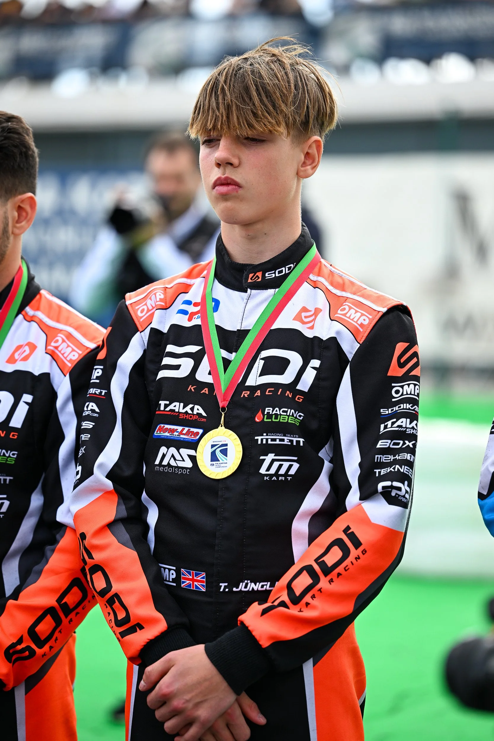 Young male race car driver wearing a black and orange racing suit with various sponsor logos, standing with a medal around his neck, on a race track, with other racers and spectators in the background.