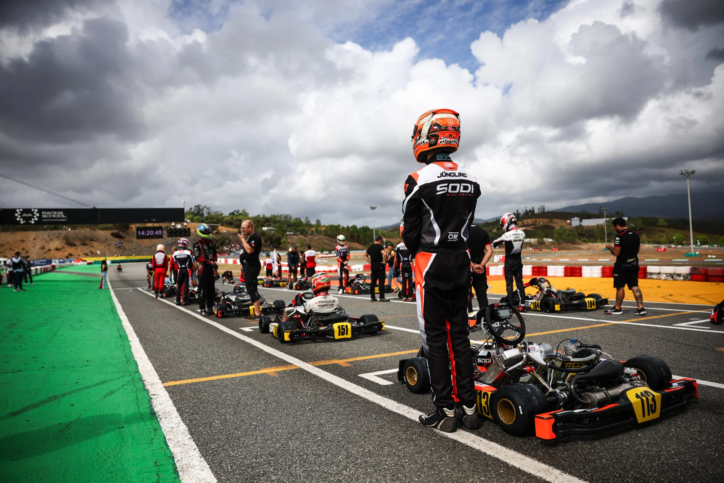 Race car drivers and team members preparing go-karts at a race track pit lane under dark cloudy skies.