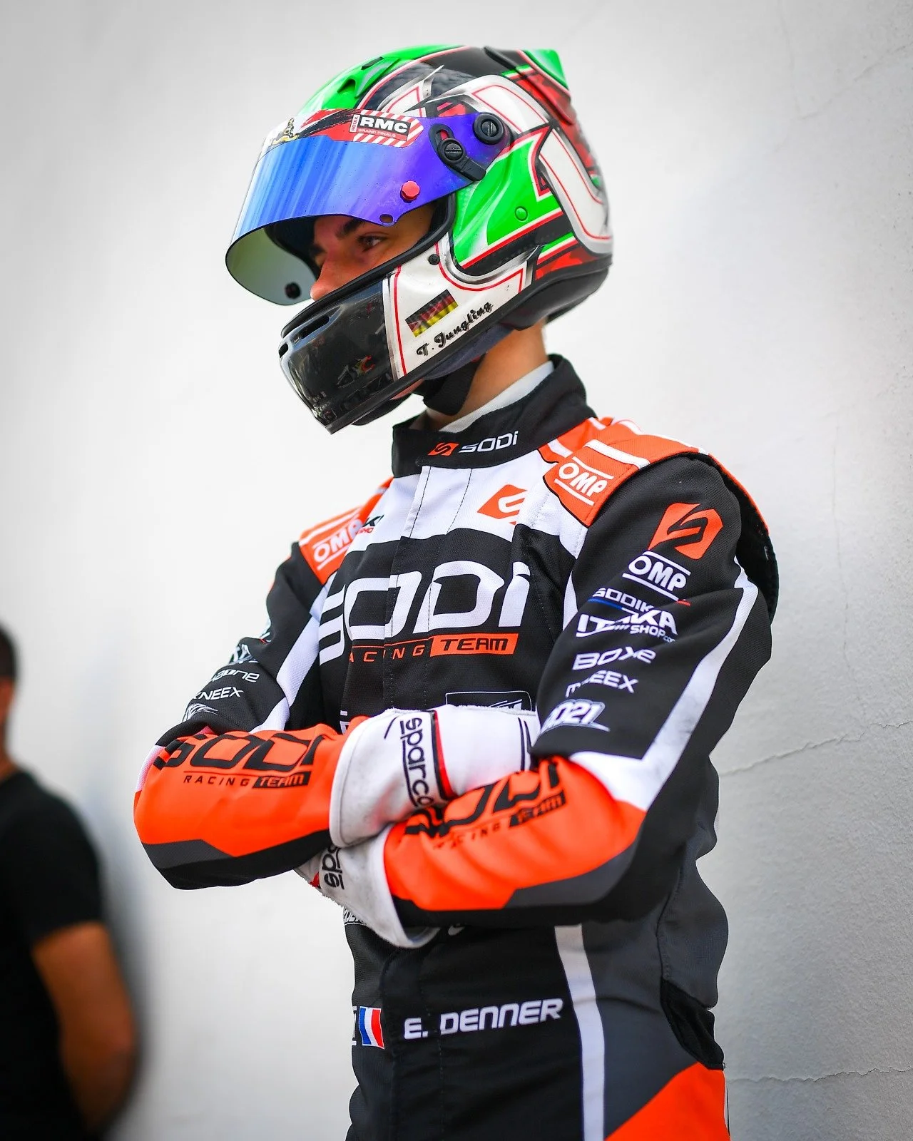 A professional race car driver wearing a helmet with green, white, red, and black colors, and a racing suit with logos and the name E. Denner.