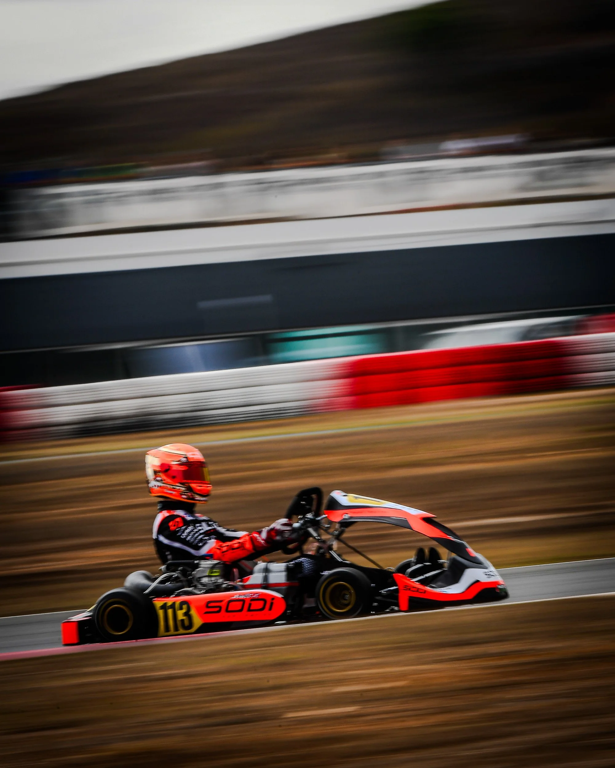 A person driving a go-kart on a race track, wearing a red helmet and racing suit, with the number 113 and the brand SODI on the kart, during daytime.