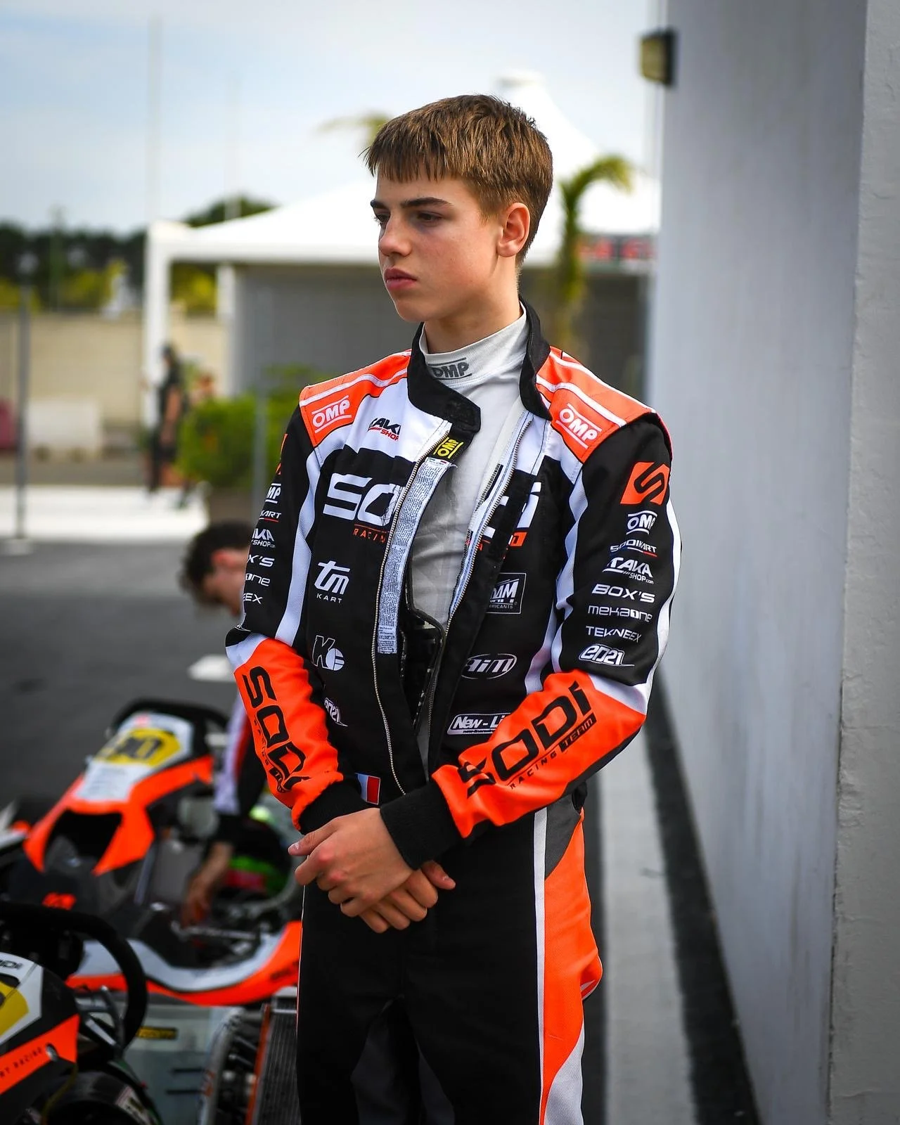 A young male racing driver in a black and orange racing suit with sponsor logos, standing outdoors near a garage and racing motorcycles.