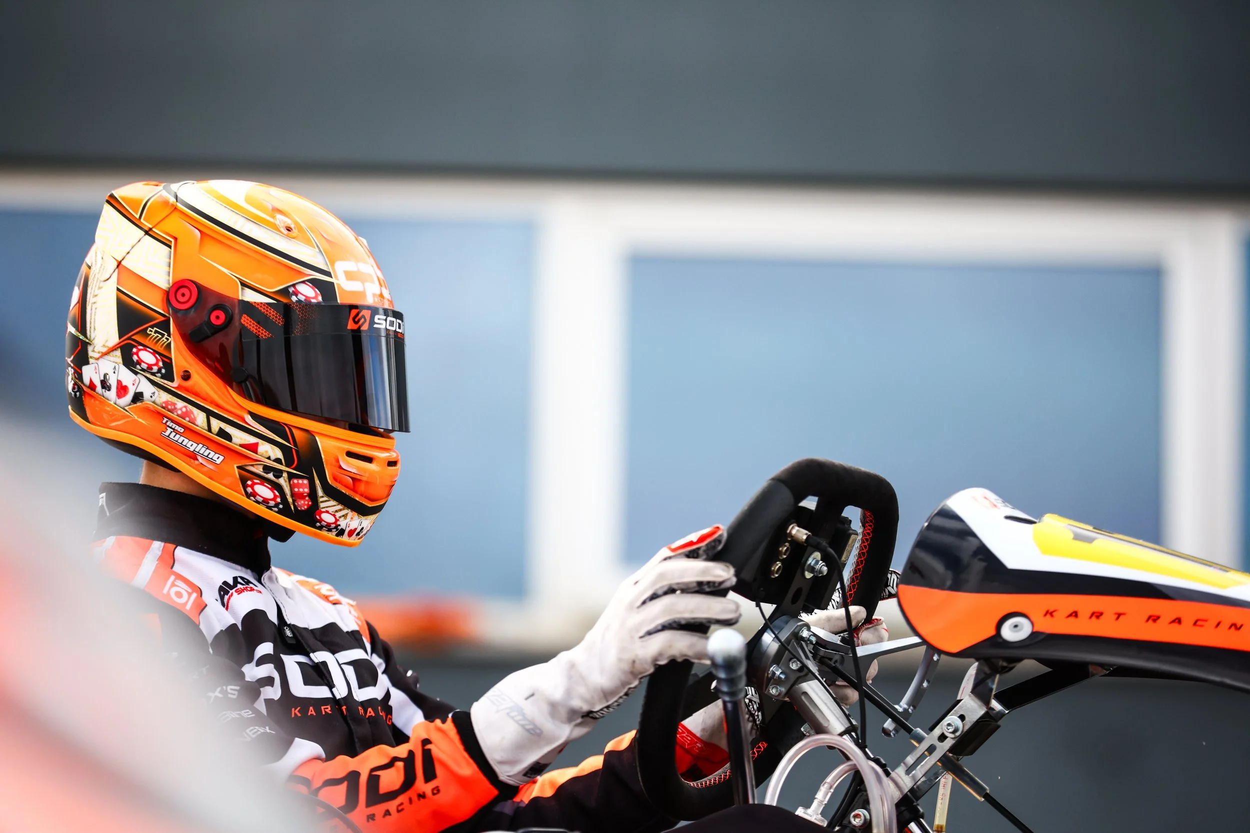 A person in racing gear and a colorful helmet sitting in a go-kart, holding a steering wheel, preparing for a race.