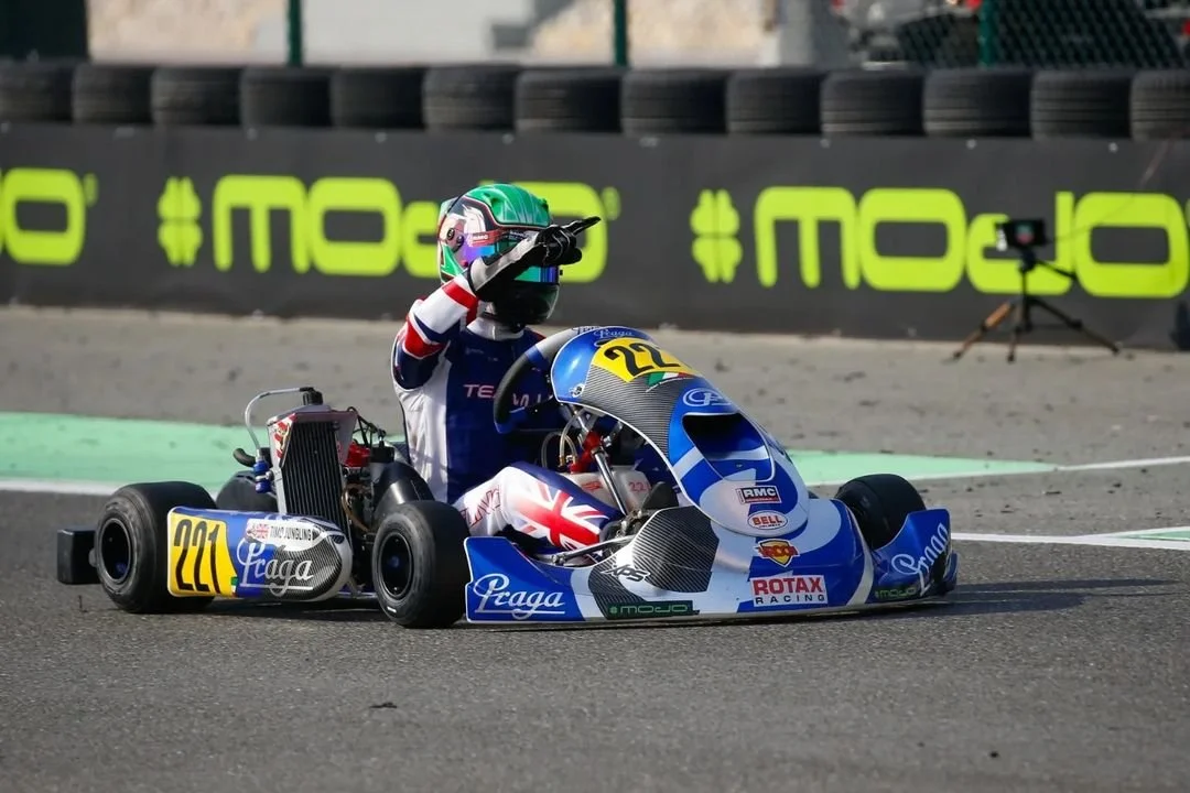 A go-kart racer wearing a helmet and racing suit sitting in a go-kart on a racetrack, gesturing with one hand.