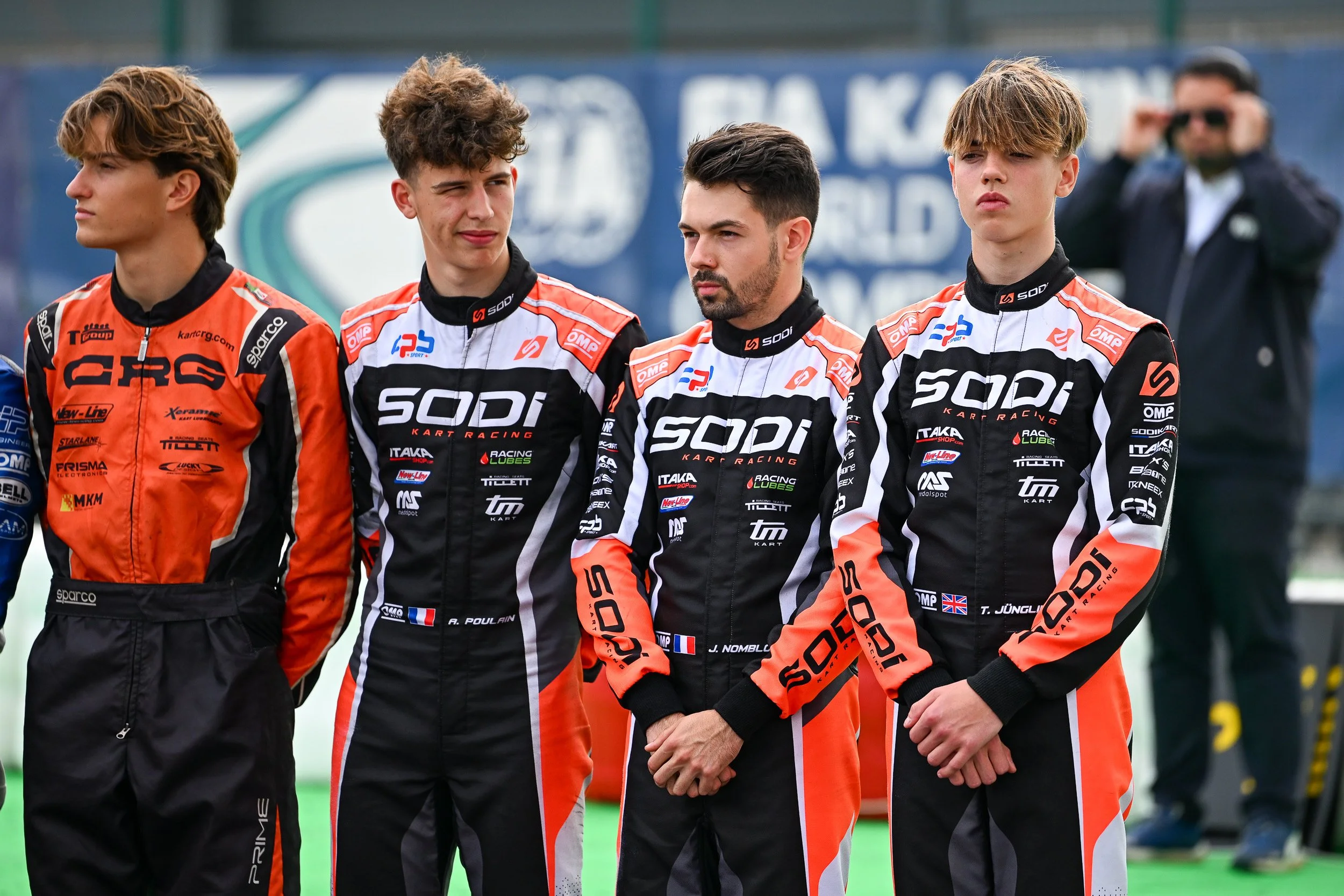 Four young men in racing suits standing in a line, with a woman in the background taking photos. They are part of a kart racing team.