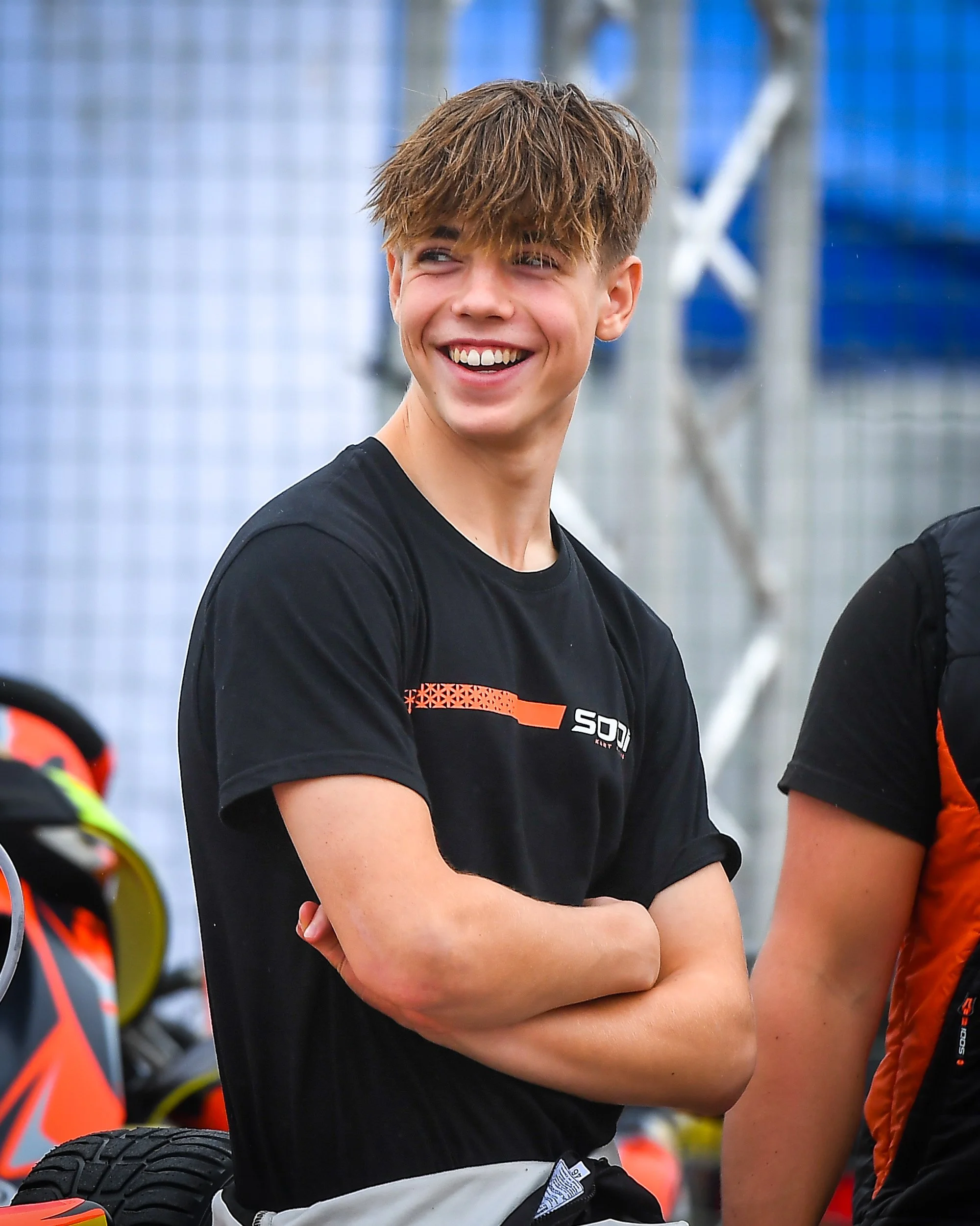 A young man smiling with crossed arms, wearing a black t-shirt, standing outdoors in a sports or racing environment.