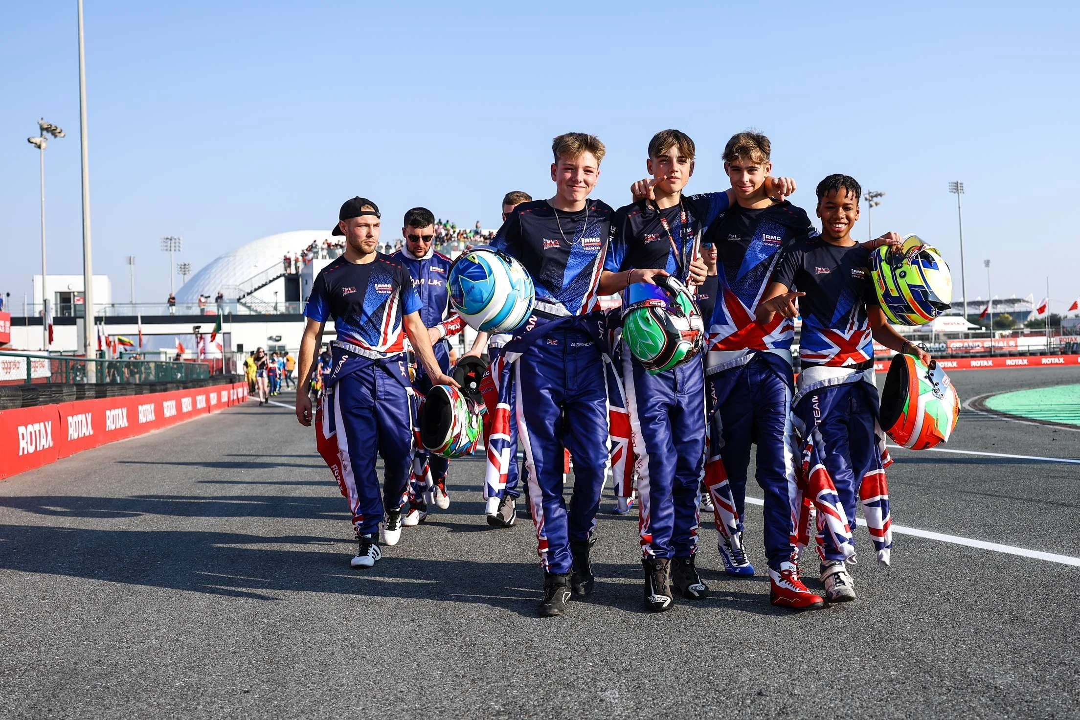 Young race car drivers wearing racing suits with Union Jack design, holding helmets, walking on race track, with grandstands and bright blue sky in background.