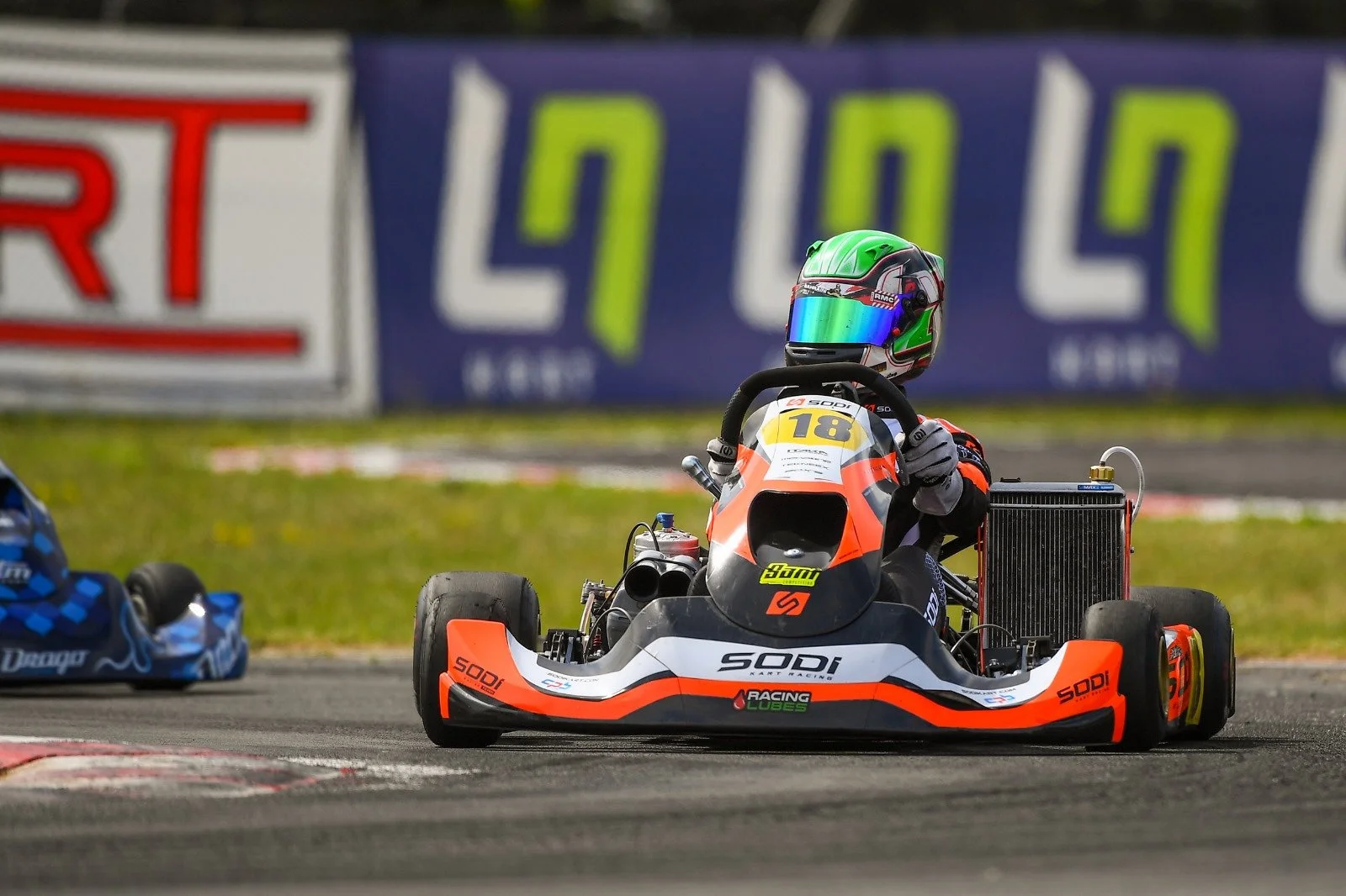 Go-kart race car on track with a driver wearing a green helmet and black racing suit, with a blue and black kart visible to the left, and a large advertisement banner in the background.