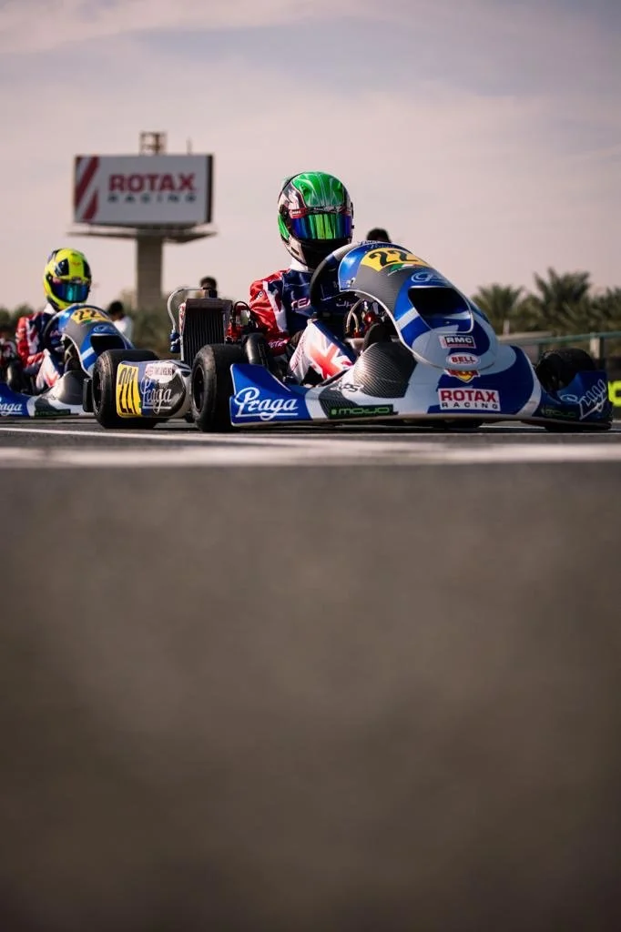 Race car drivers in blue and white go-karts on a race track, with one driver in a green helmet and others in yellow helmets, under a cloudy sky with a billboard in the background displaying 'ROTAX RACING'.