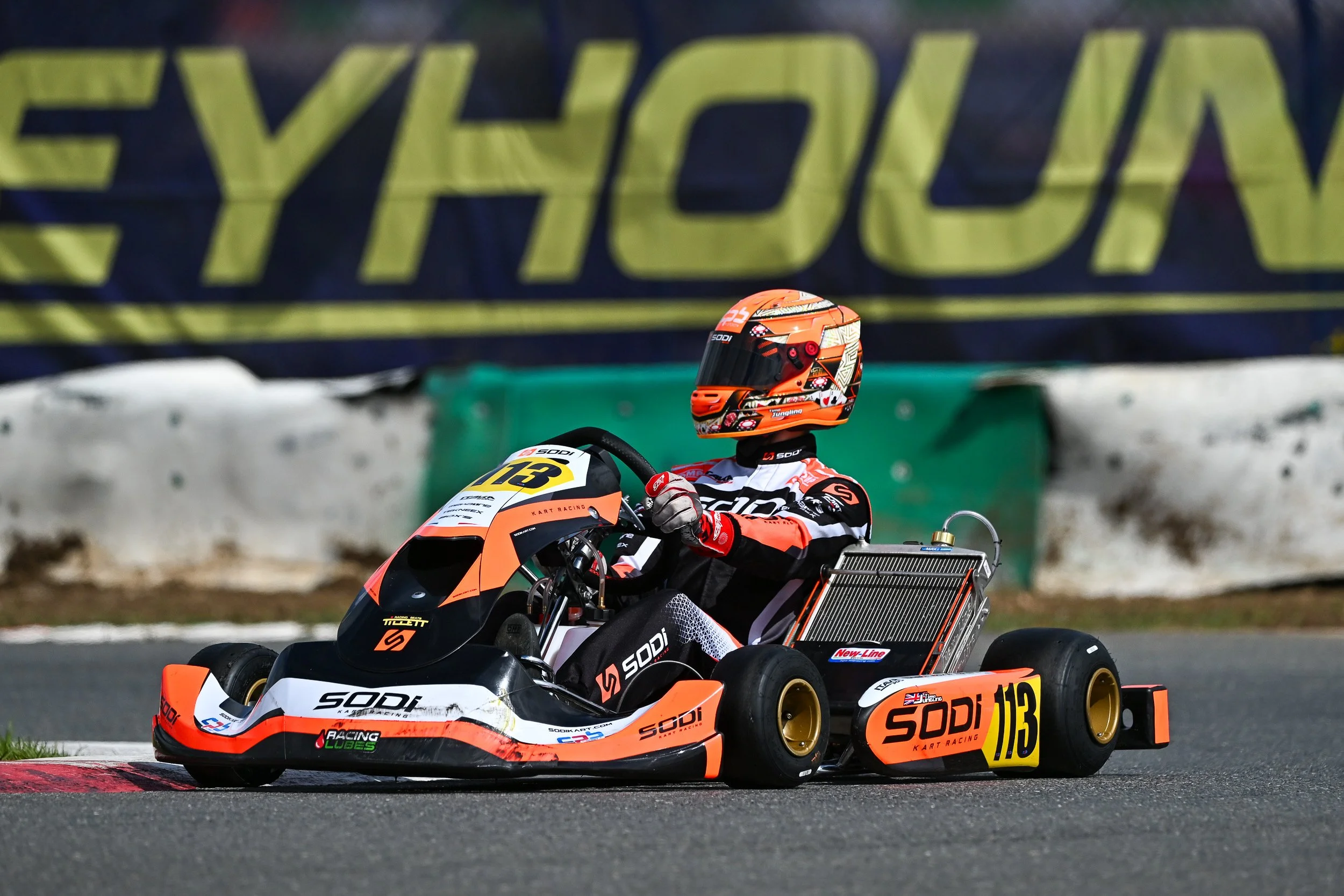 A go-kart racer wearing a helmet and racing suit driving on a race track with a large banner in the background.