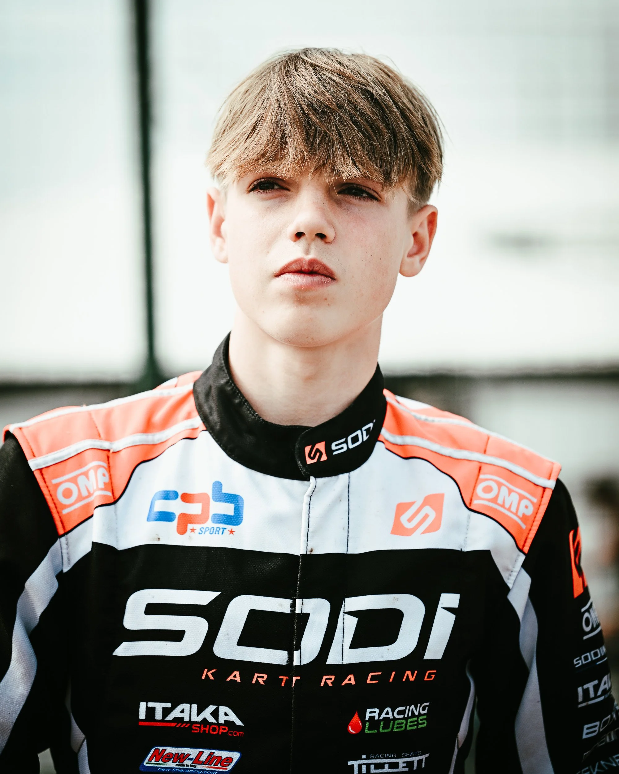 A young male race car driver in a black and orange racing suit with sponsor logos, looking pensively to the side.