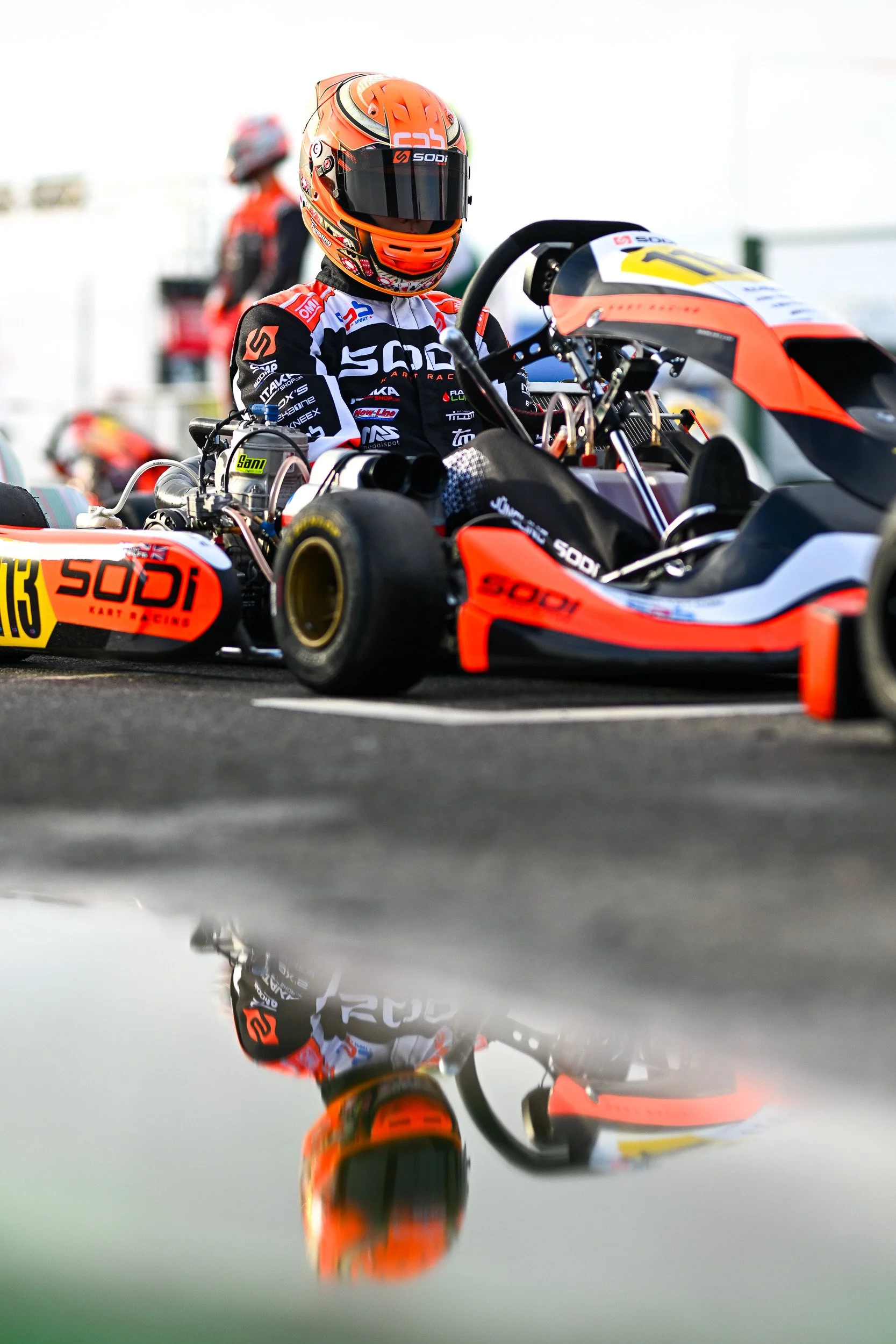 Race car driver in orange helmet and black racing suit sitting in a go-kart on track, with reflection of go-kart and driver in a puddle on the ground.
