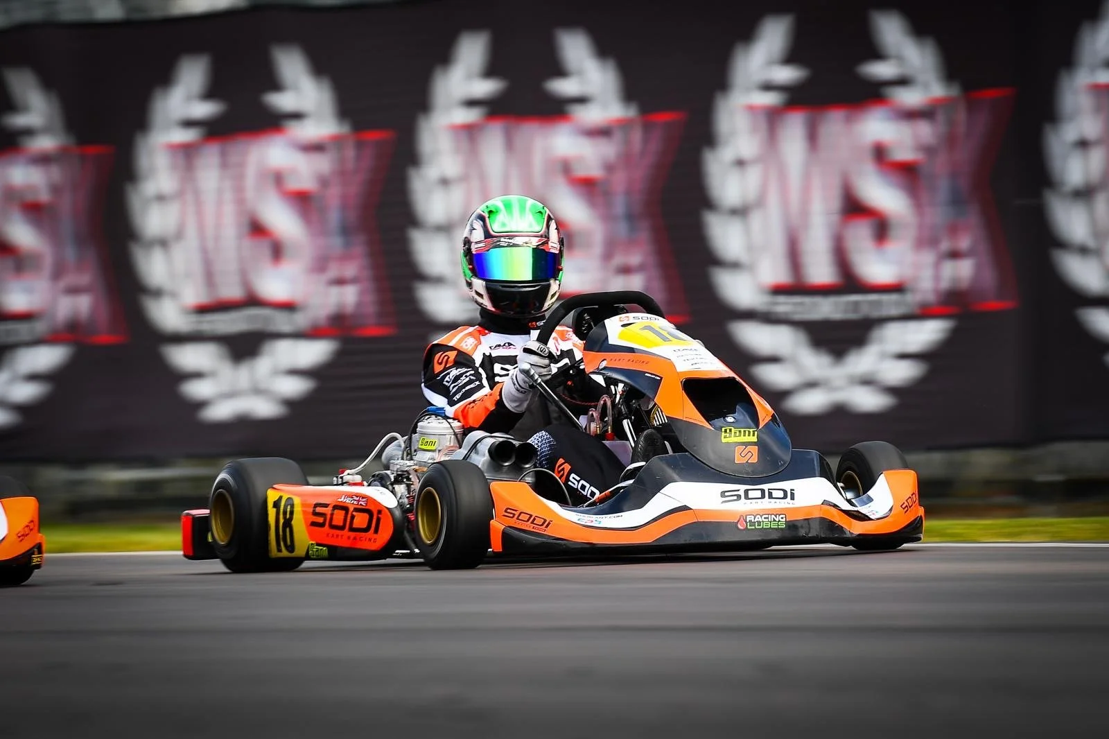 Race car driver in orange and black racing suit driving a go-kart on a track, with a blurred background featuring logos.