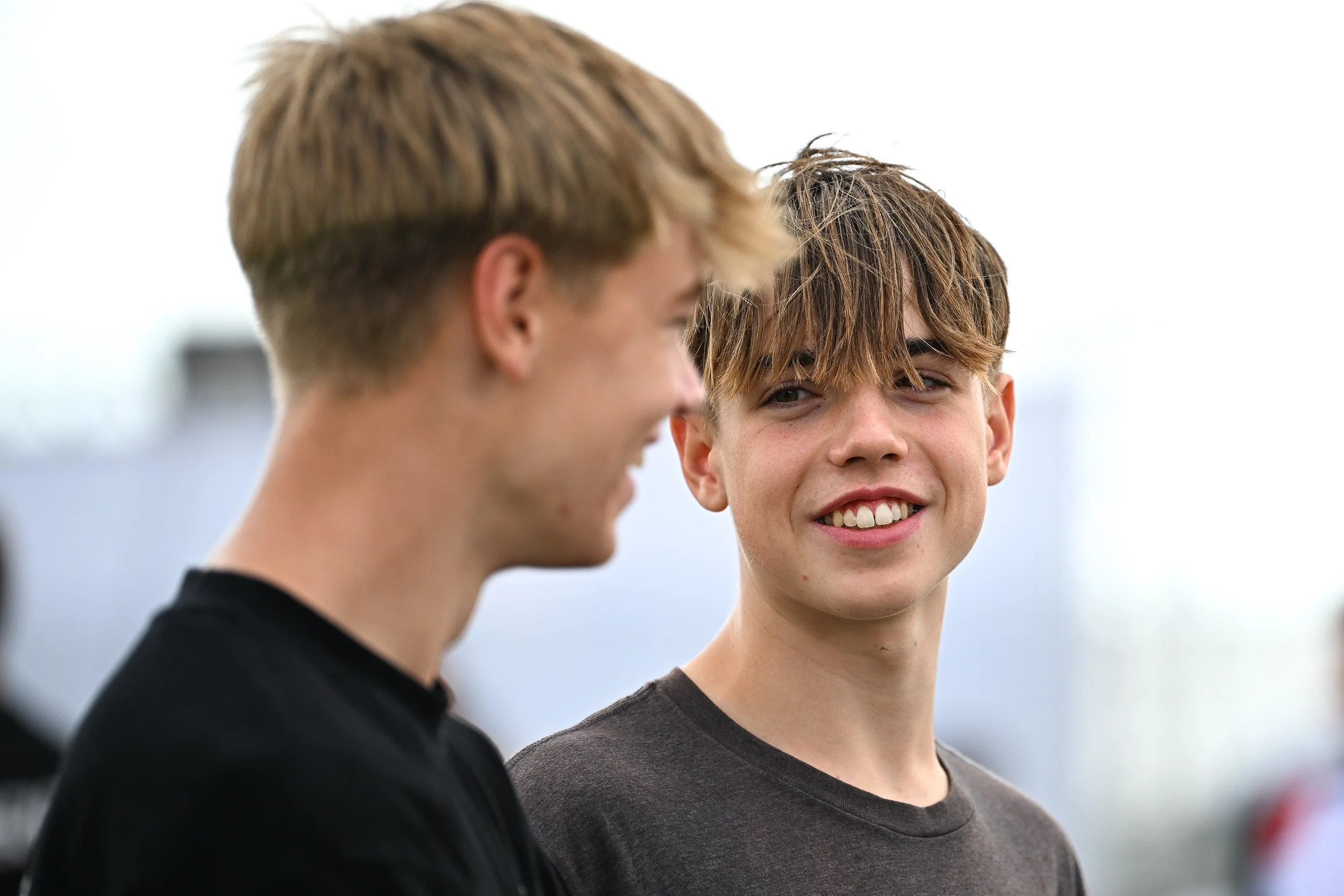 Two young men smiling and talking outdoors, one facing camera and the other with back partially turned.
