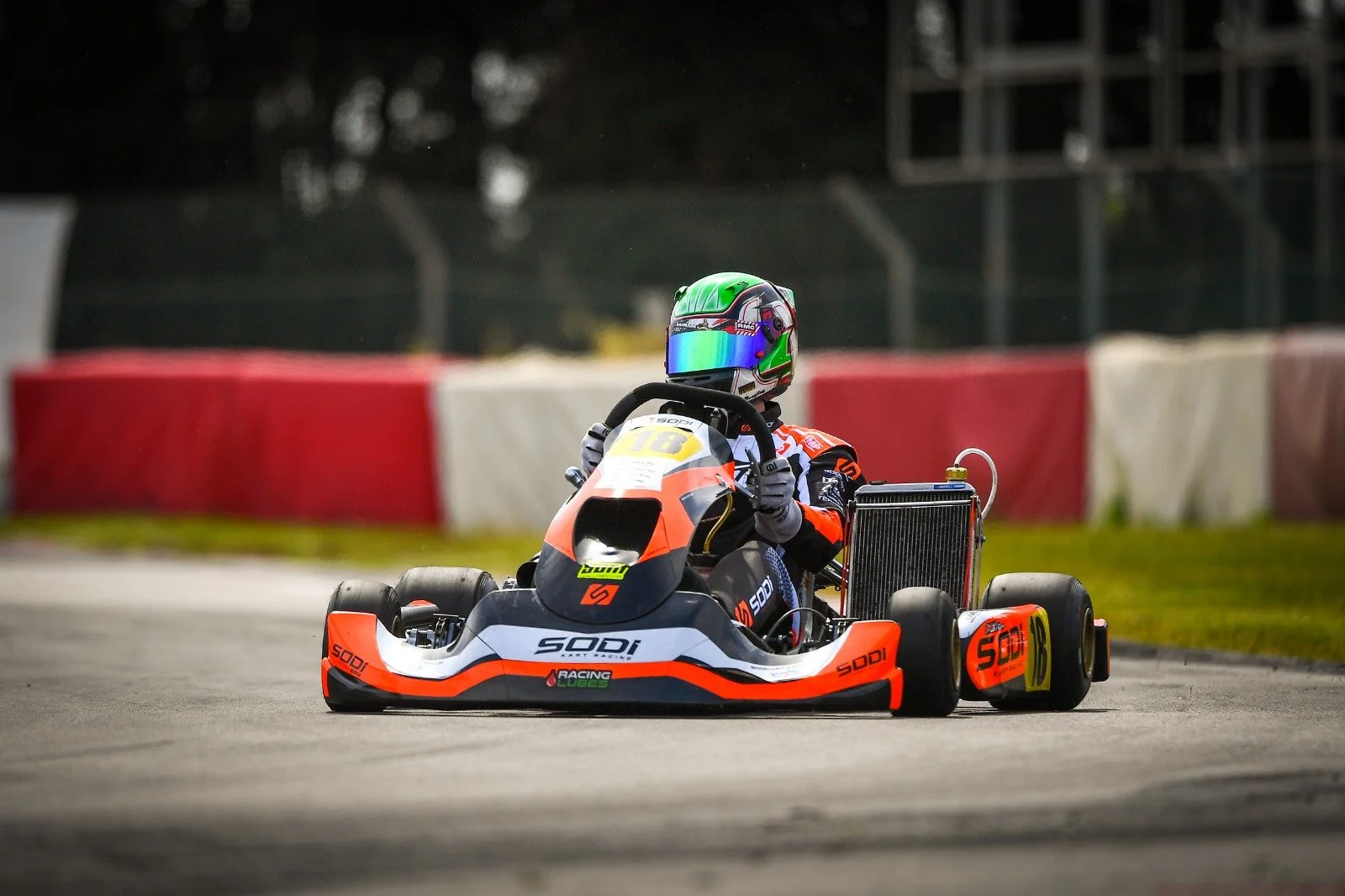 Go-kart racer wearing a colorful helmet and racing suit driving a go-kart on a racetrack with red and white barriers in the background.