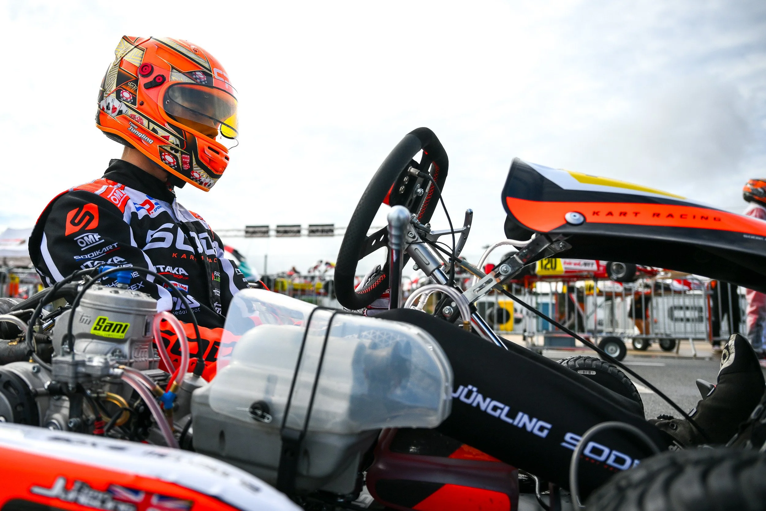 A race car driver in a helmet and racing suit sitting in a go-kart on a racing track, with others and equipment visible in the background.