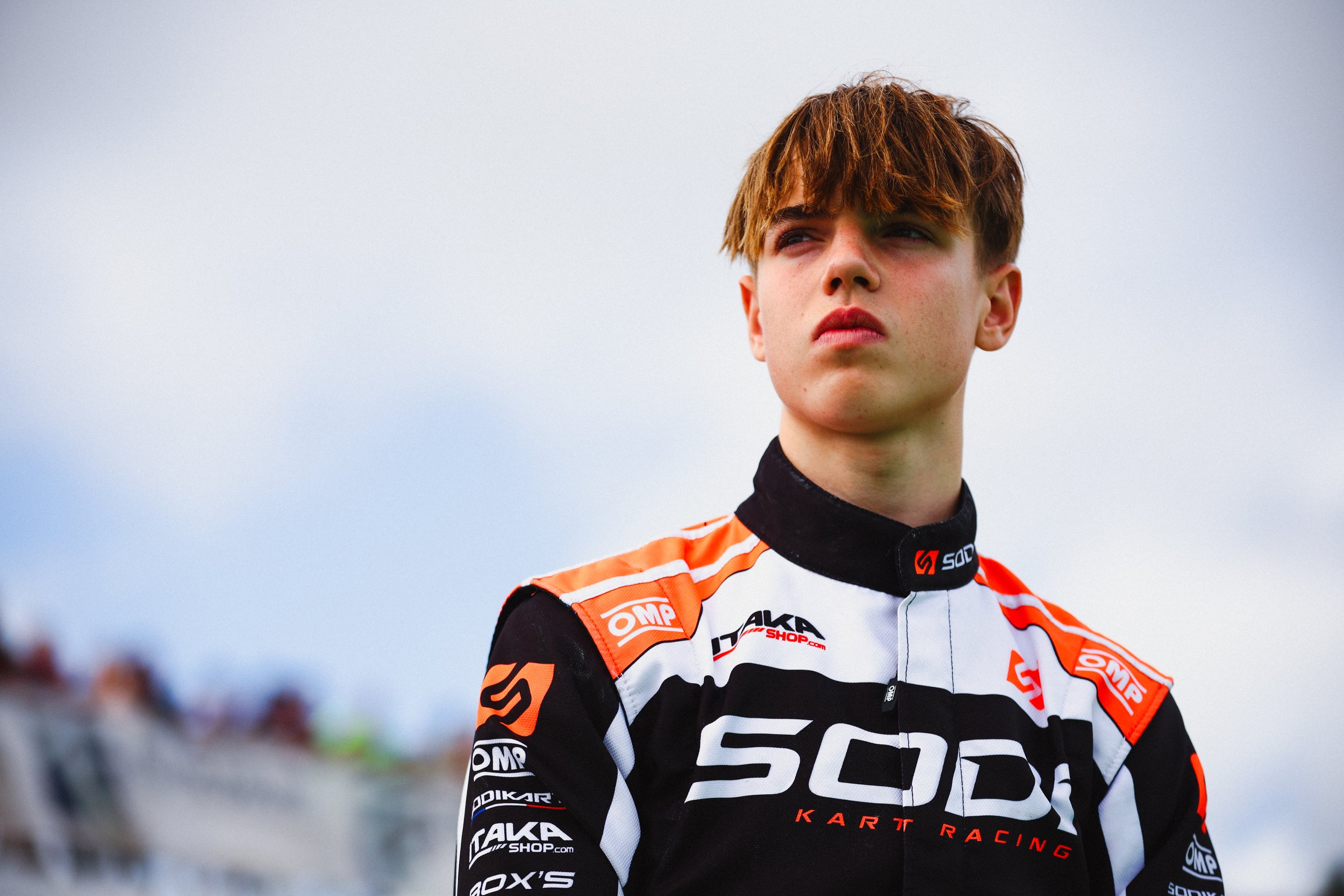 Young male race car driver in black, white, and orange racing suit with sponsor logos standing outdoors, looking to the side with a serious expression, against a cloudy sky background.