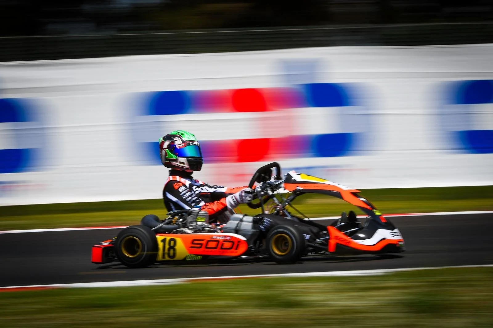 A go-kart driver wearing a colorful helmet driving on a race track with motion blur in the background.