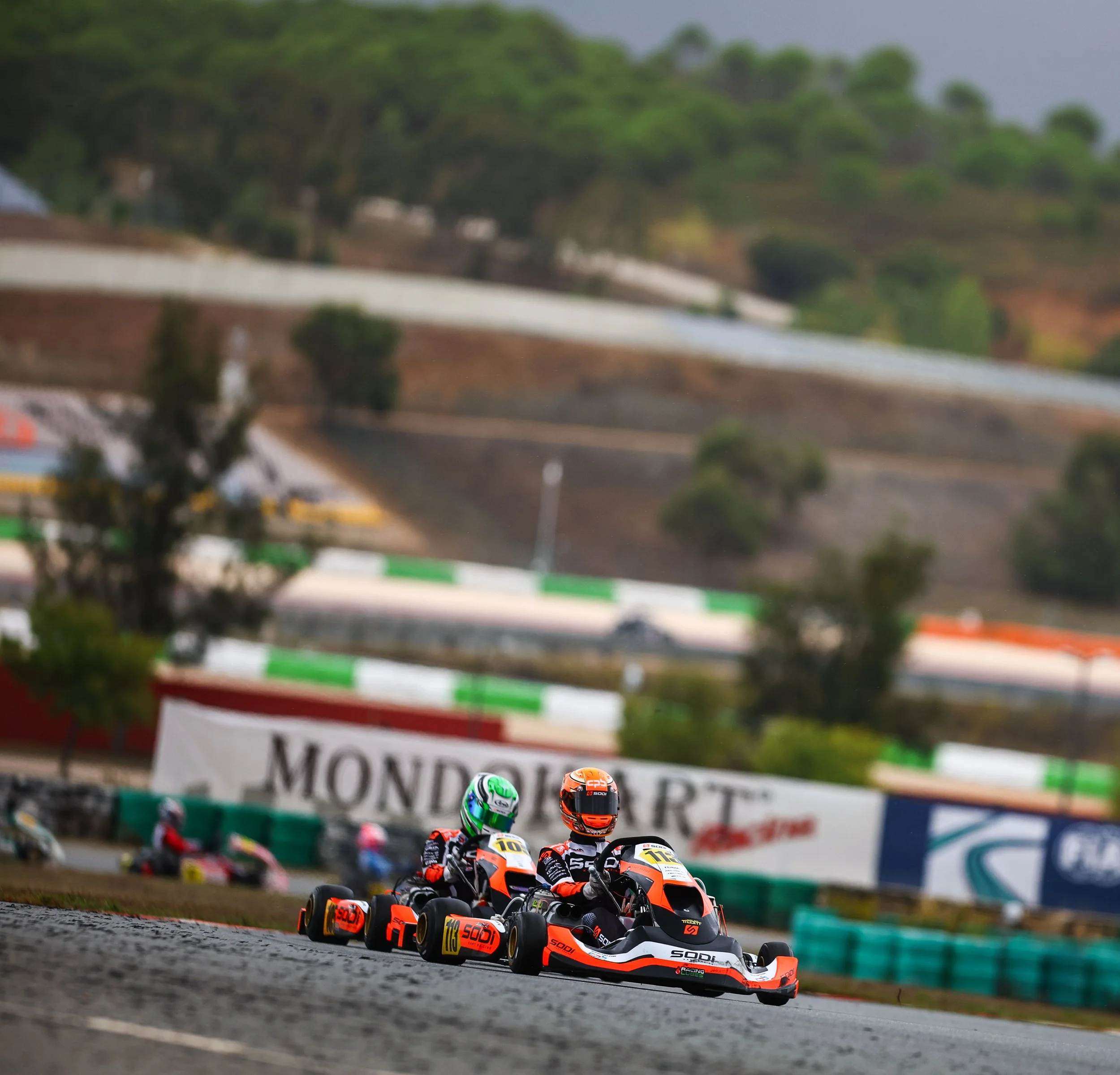 Two go-kart racers in black and orange suits and helmets speeding on a race track, with a scenic backdrop of hills and trees.