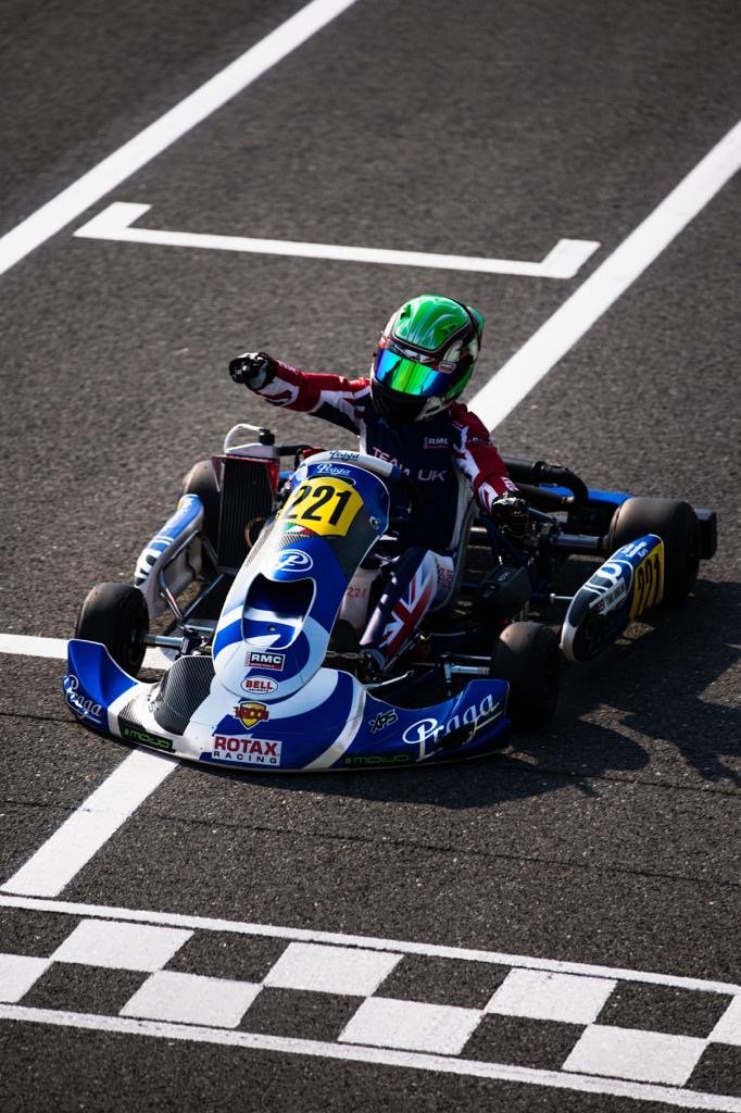 Go-kart racer celebrating crossing the finish line on a racetrack, wearing a green helmet and racing suit with Union Jack flag design, blue and white go-kart with number 221 and sponsor decals.
