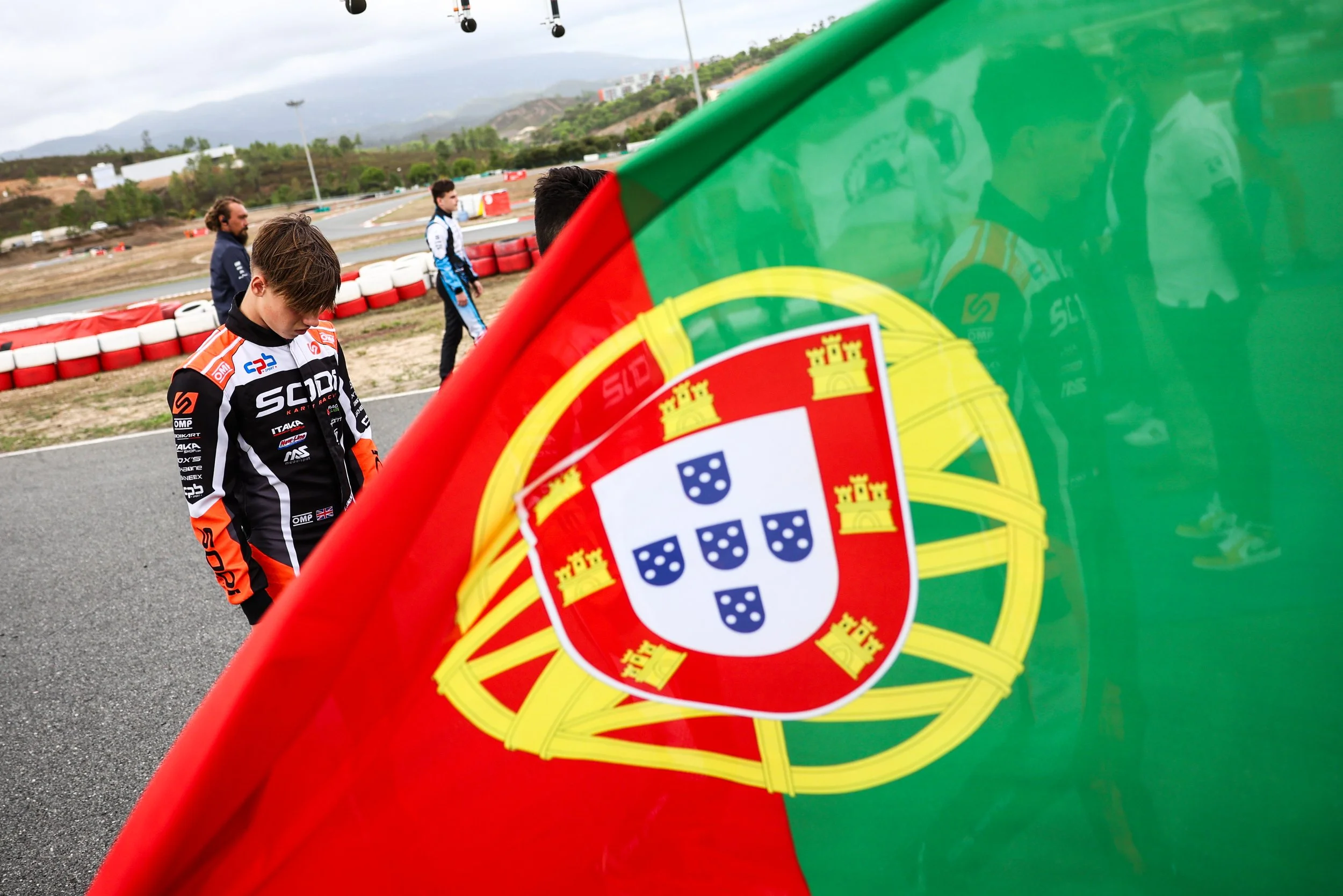 Close-up of a Portuguese flag with a racing driver and others in the background at a race track.