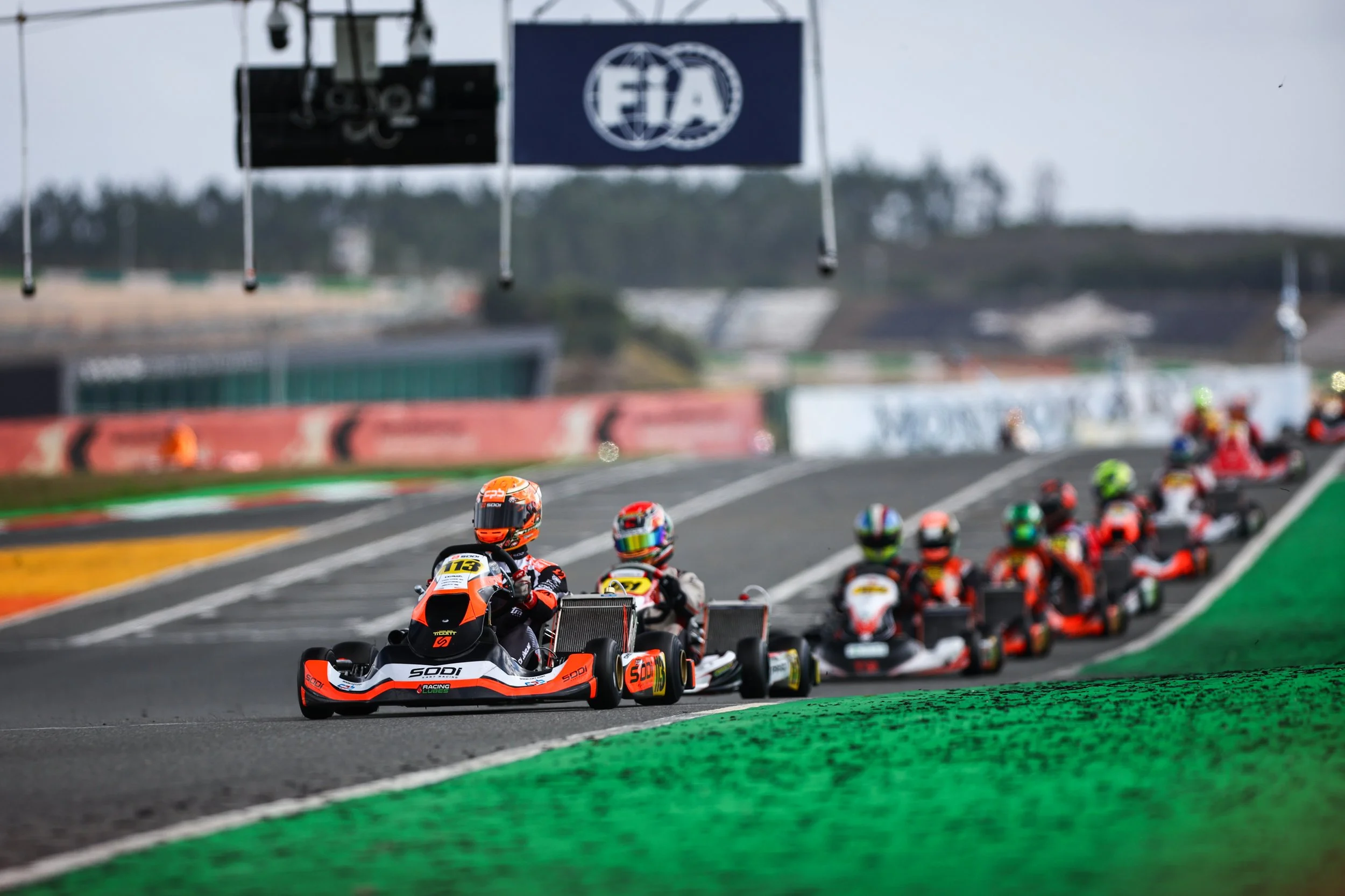 Go-kart race on a paved track with multiple drivers in colorful helmets and suits, under a sign with FIA logo.
