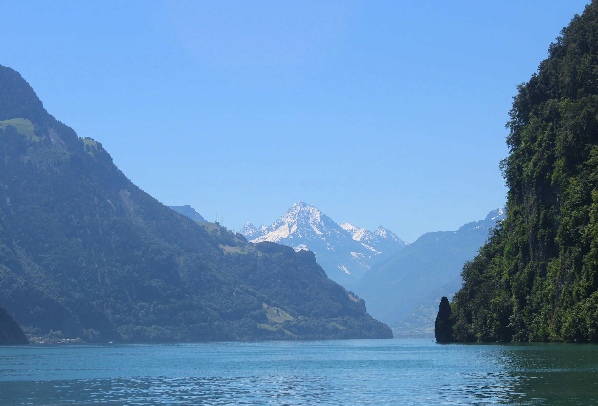 A scenic landscape featuring a lake surrounded by green mountains with snow-capped peaks under a clear blue sky.